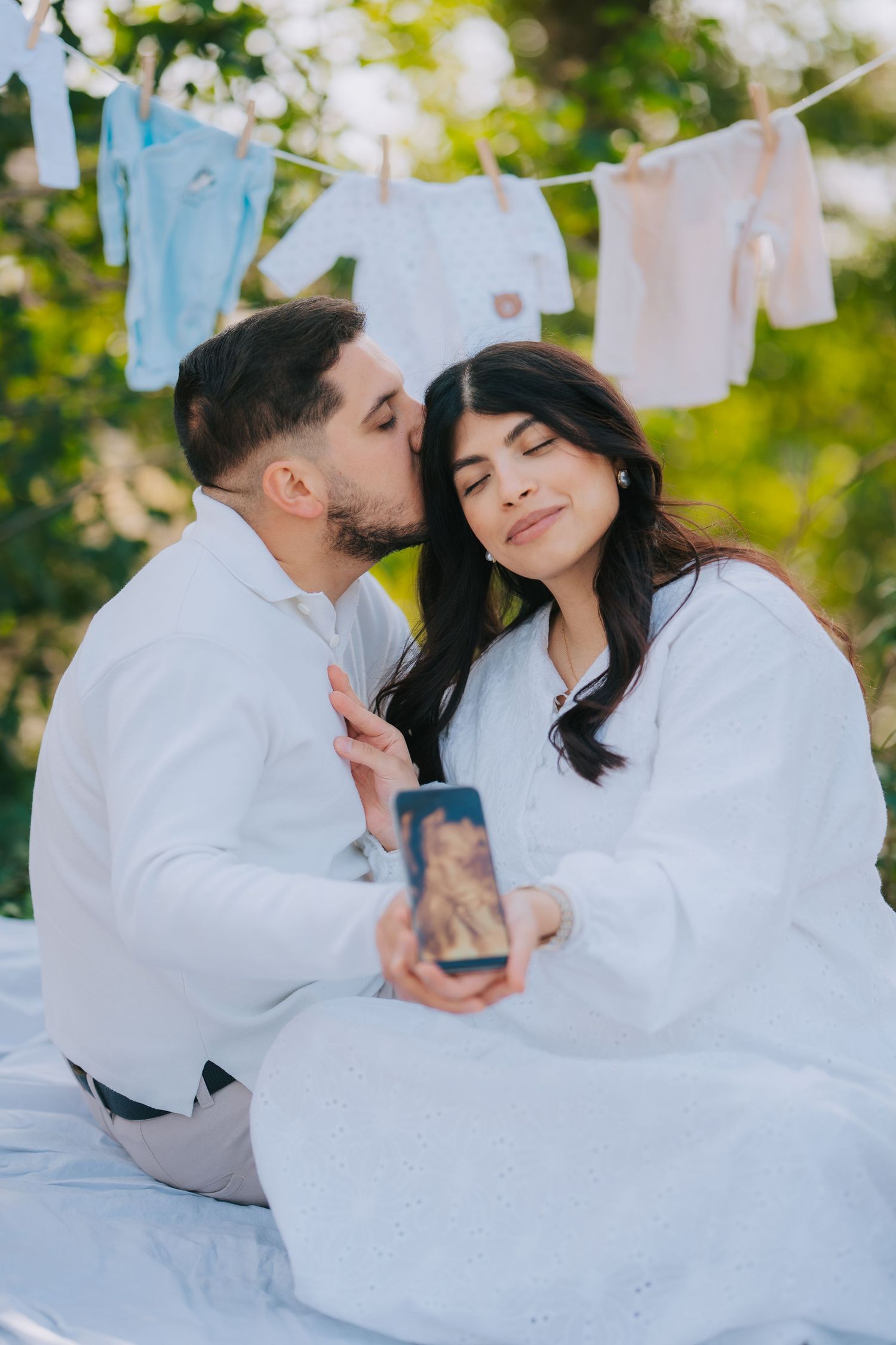 A couple in white outfits sharing an intimate moment with baby clothes hanging on a clothesline behind them.