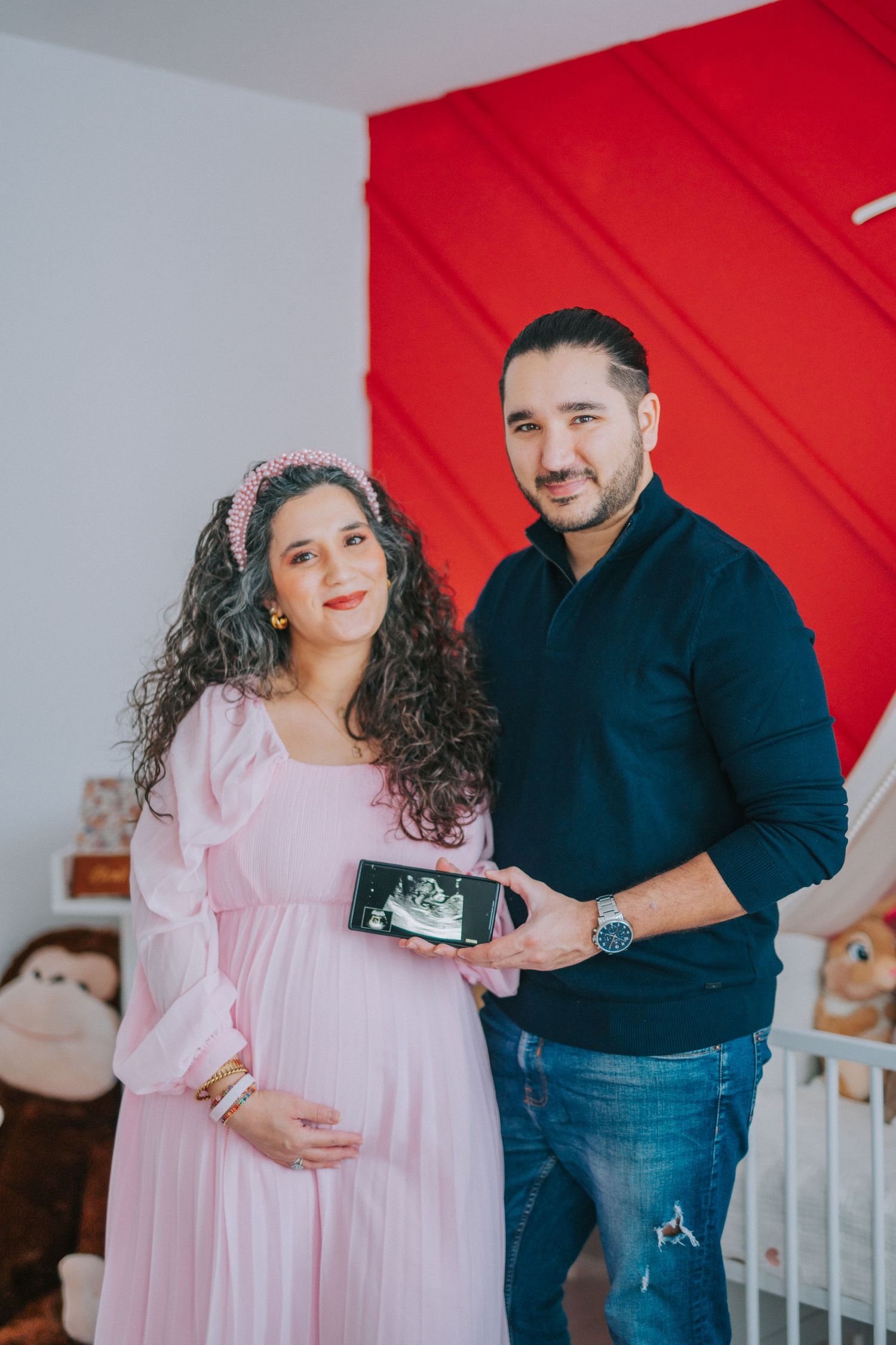Expectant couple in nursery holding ultrasound photo against red accent wall.