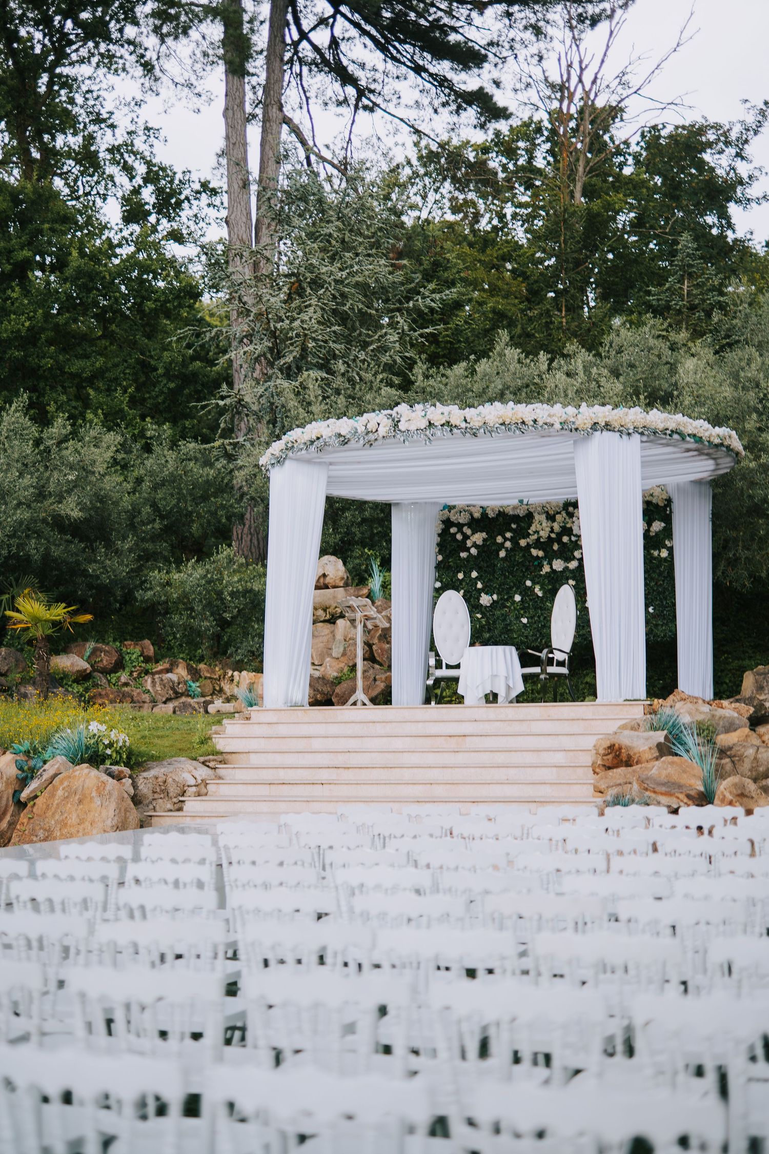 White wedding gazebo decorated with flowers overlooking rows of white chairs in garden setting.
