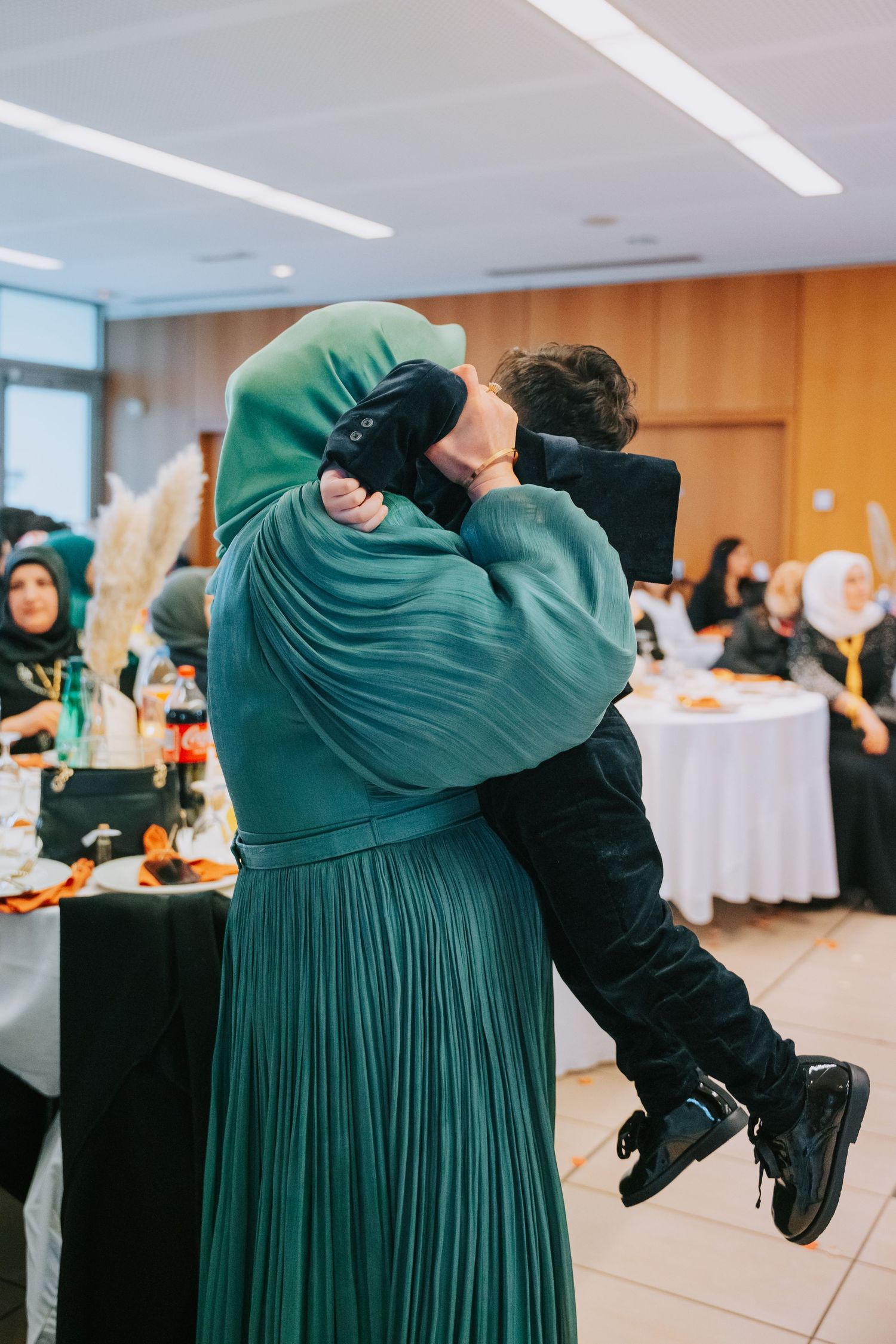 Woman in a green pleated dress holding a child at an indoor celebration.