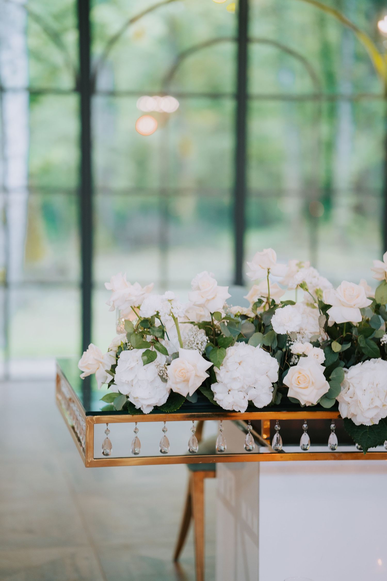 Elegant white roses and peonies arranged on a gold mirrored tray in a sunlit conservatory setting.