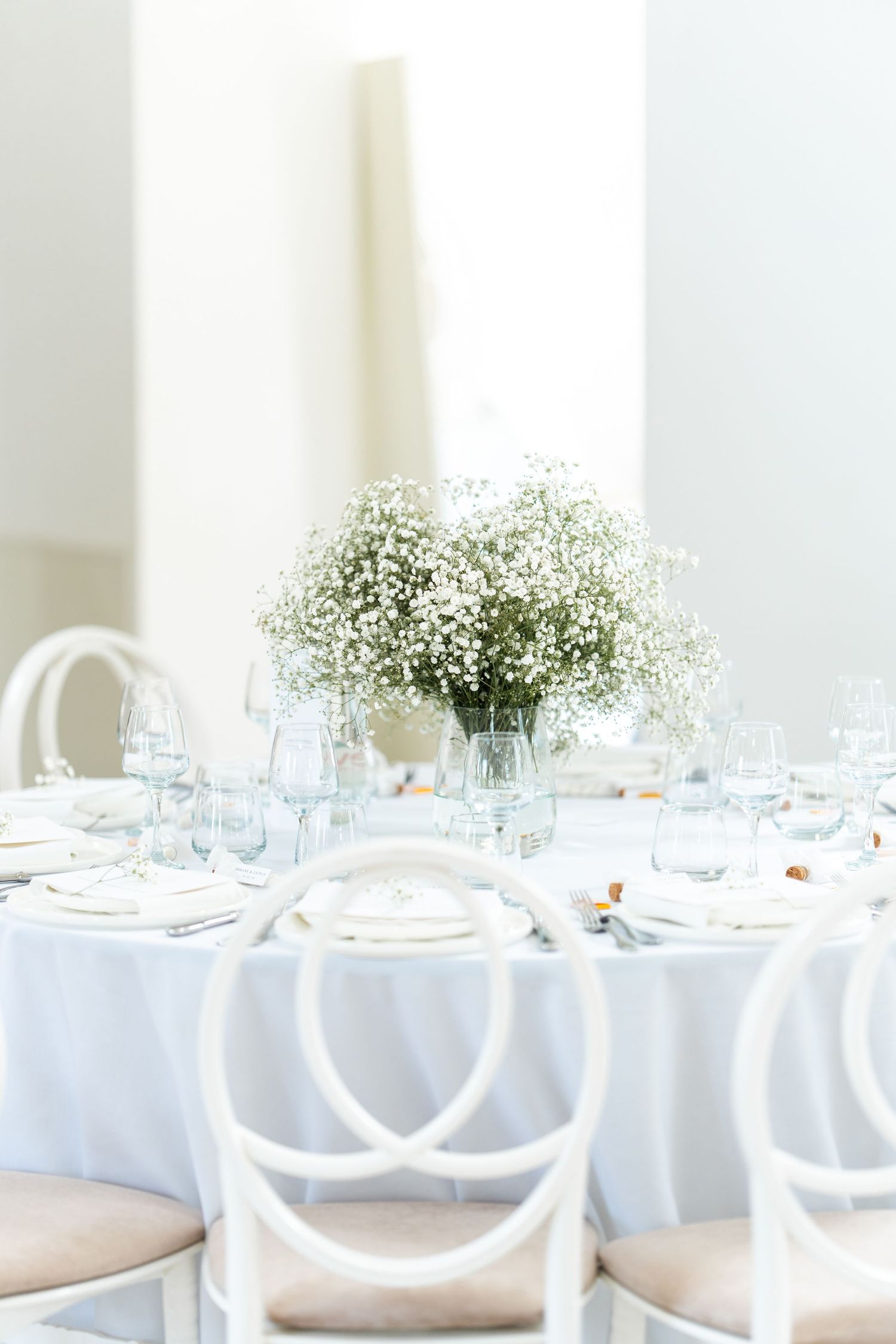 White minimalist wedding reception table setting with baby's breath centerpiece and cross-back chairs.