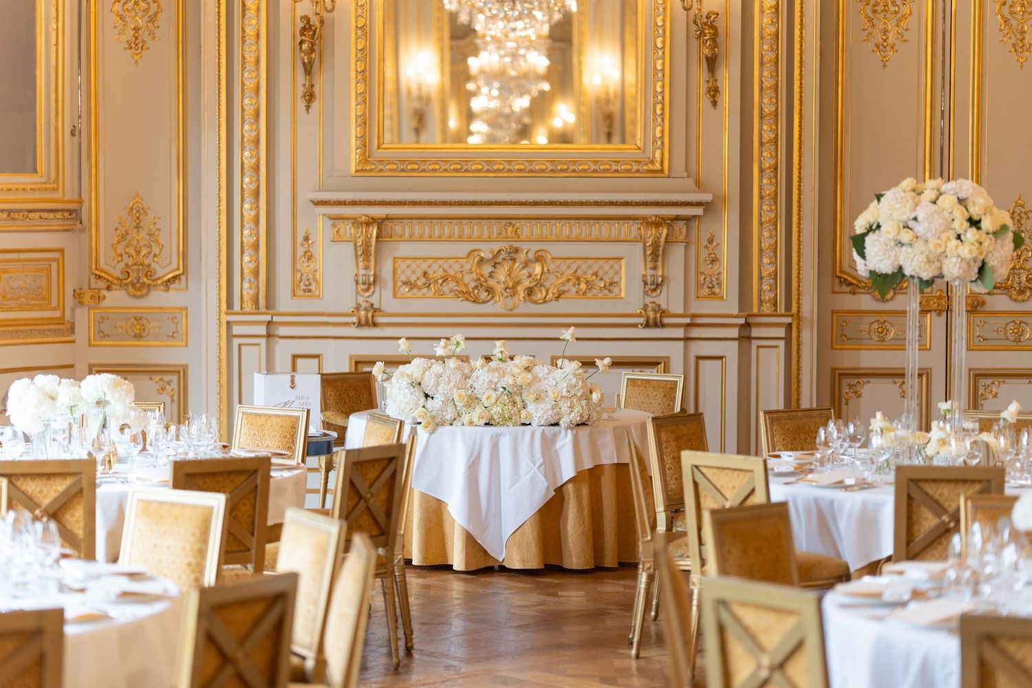 Elegant ballroom with ornate gold decor, crystal chandeliers, and round dining tables set with white linens and floral centerpieces.