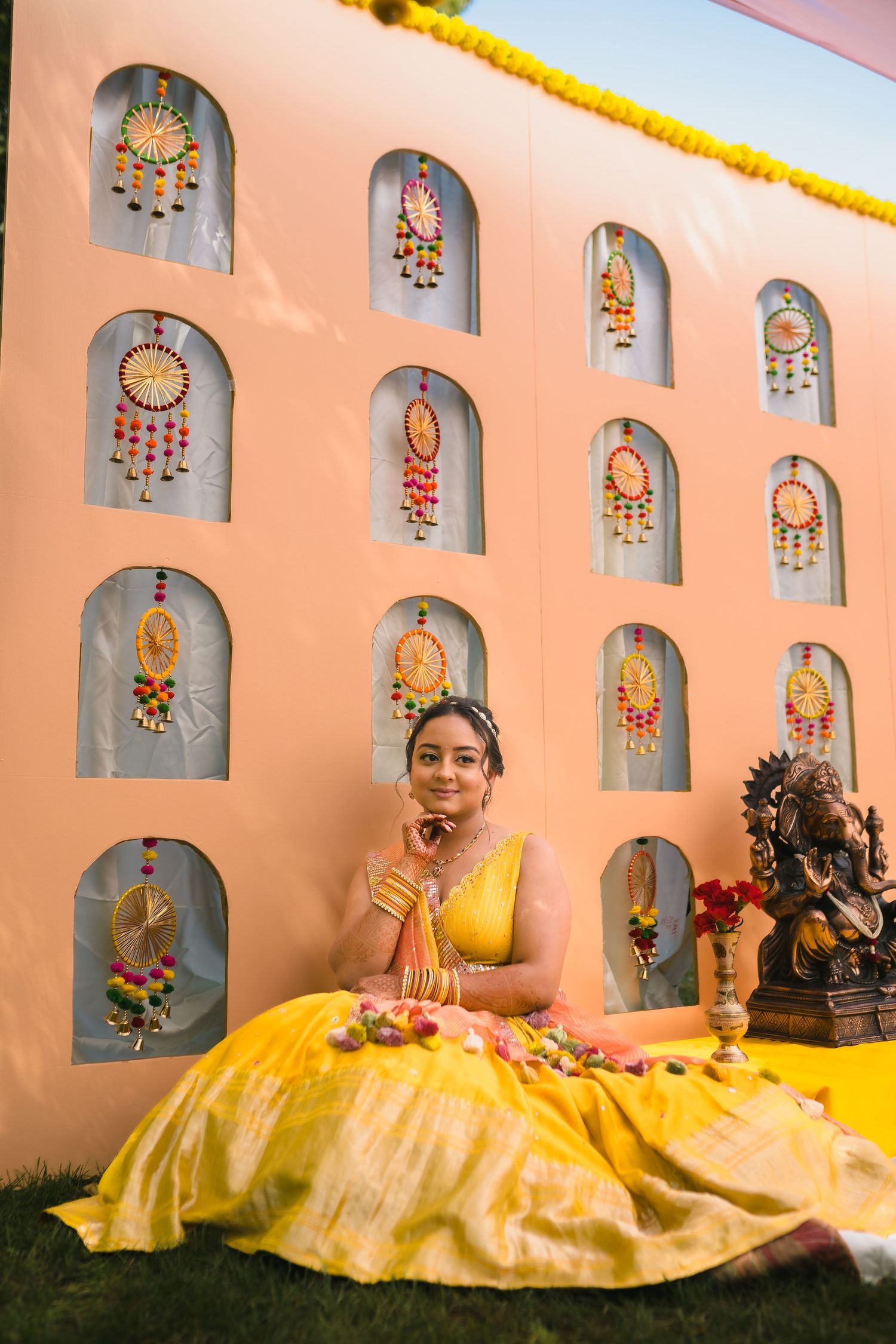 Woman in yellow lehenga sits against pink wall with decorative niches and hanging ornaments.