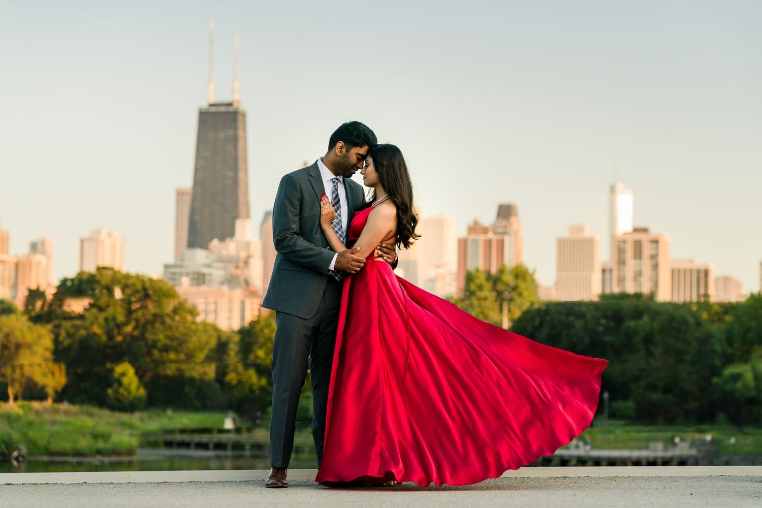 A couple in formal attire embracing against the Chicago skyline at sunset, with the bride in a flowing red gown.