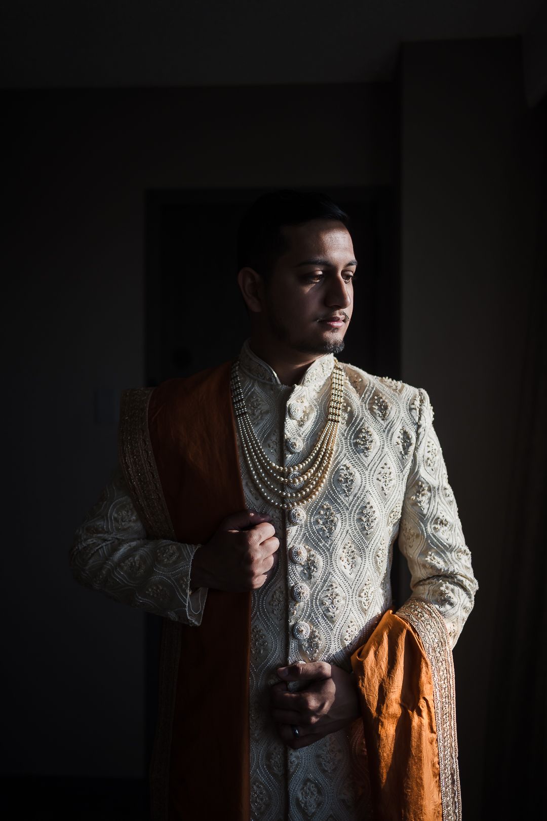 Traditional South Asian groom wearing ornate cream and gold sherwani with pearl necklaces in dramatic lighting.