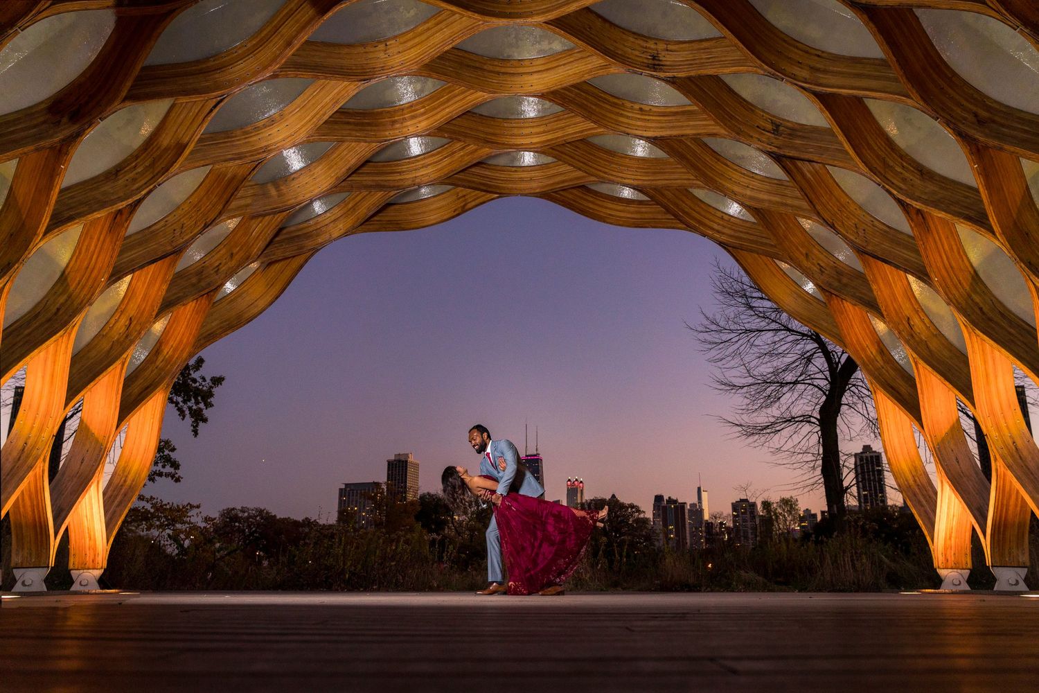 A romantic silhouette under a honeycomb wooden structure with city skyline backdrop at dusk.