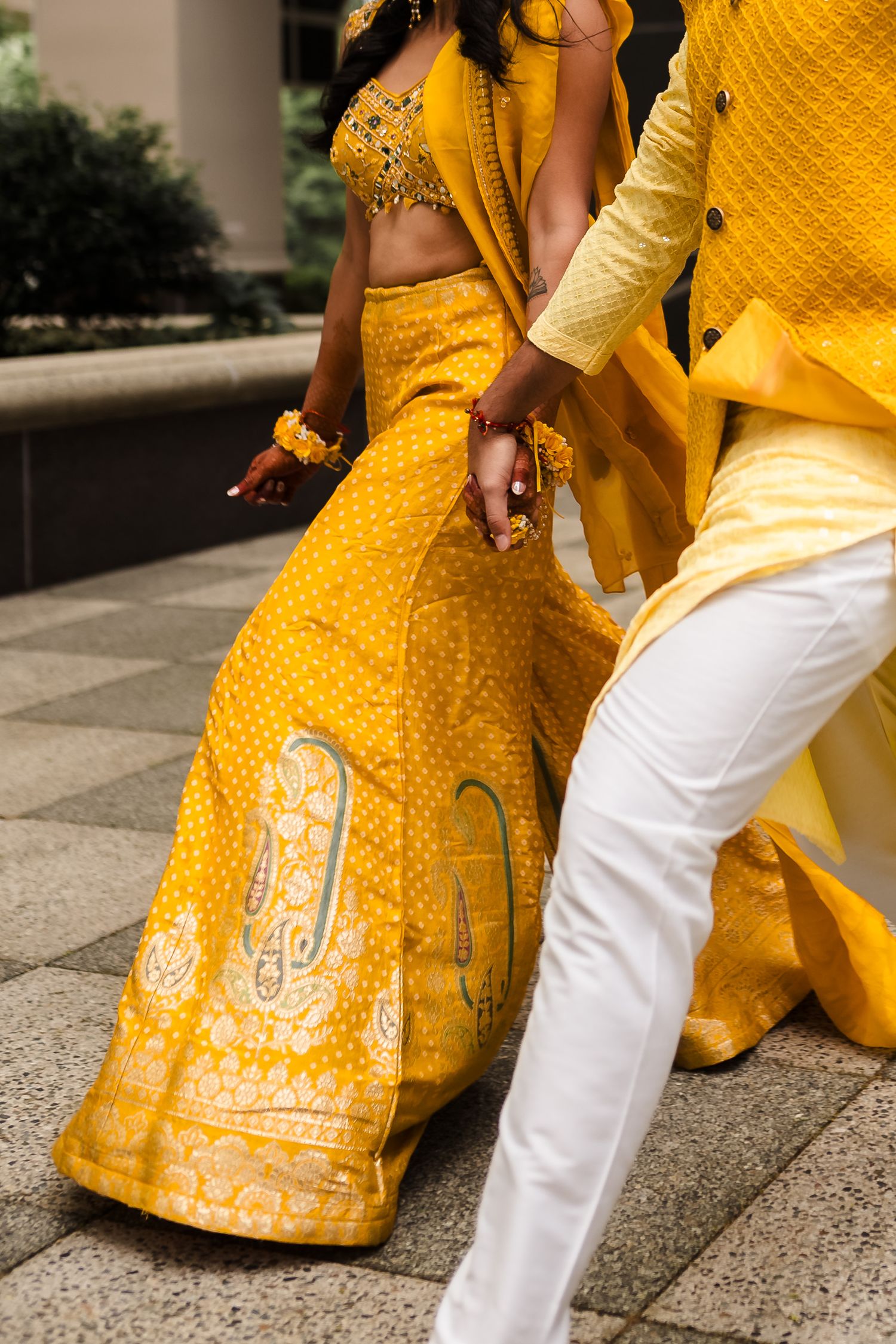 Couple in traditional yellow and white Indian wedding attire holding hands and walking on stone paving.