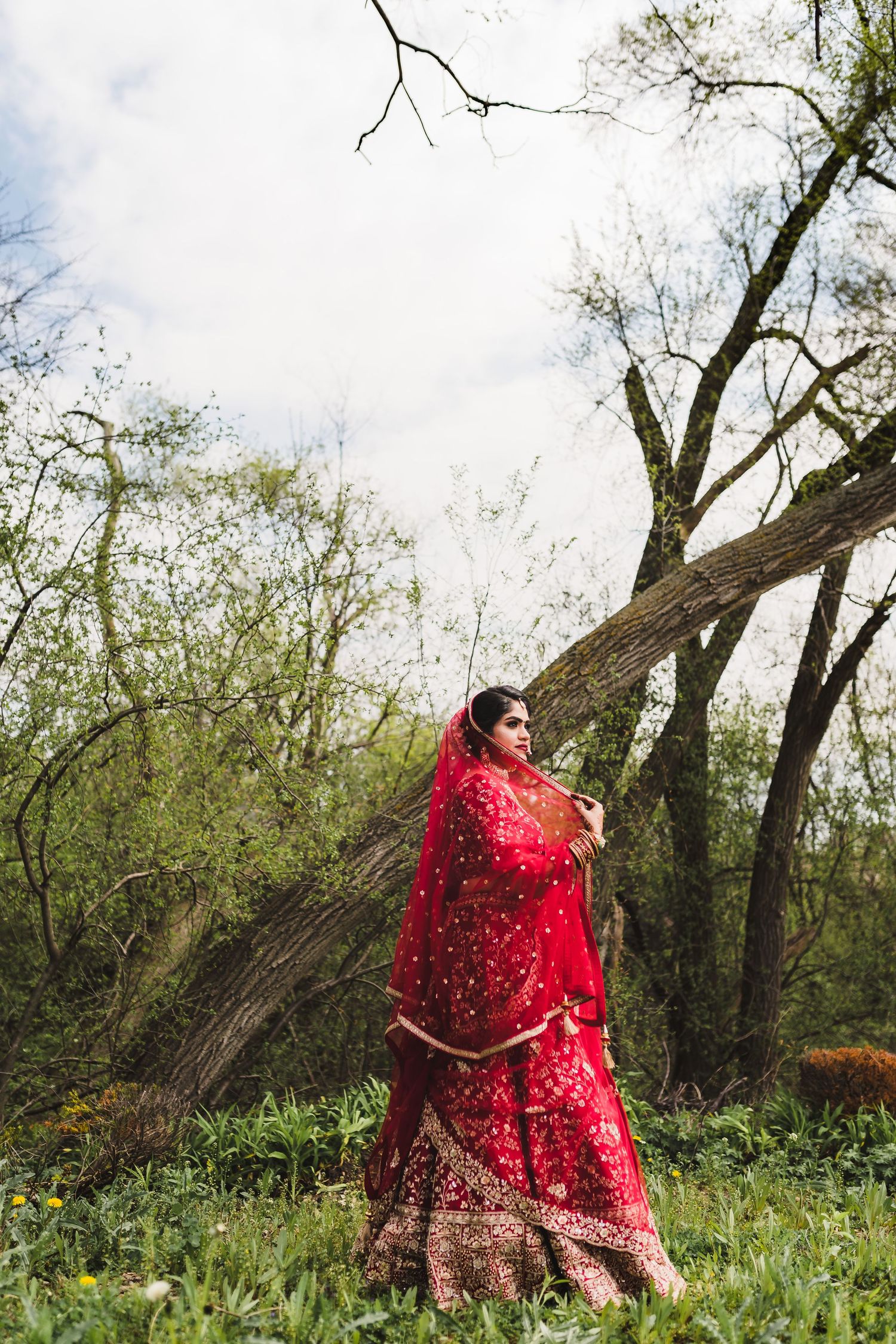 A traditional Indian bride in a red embroidered lehenga poses in a natural woodland setting.