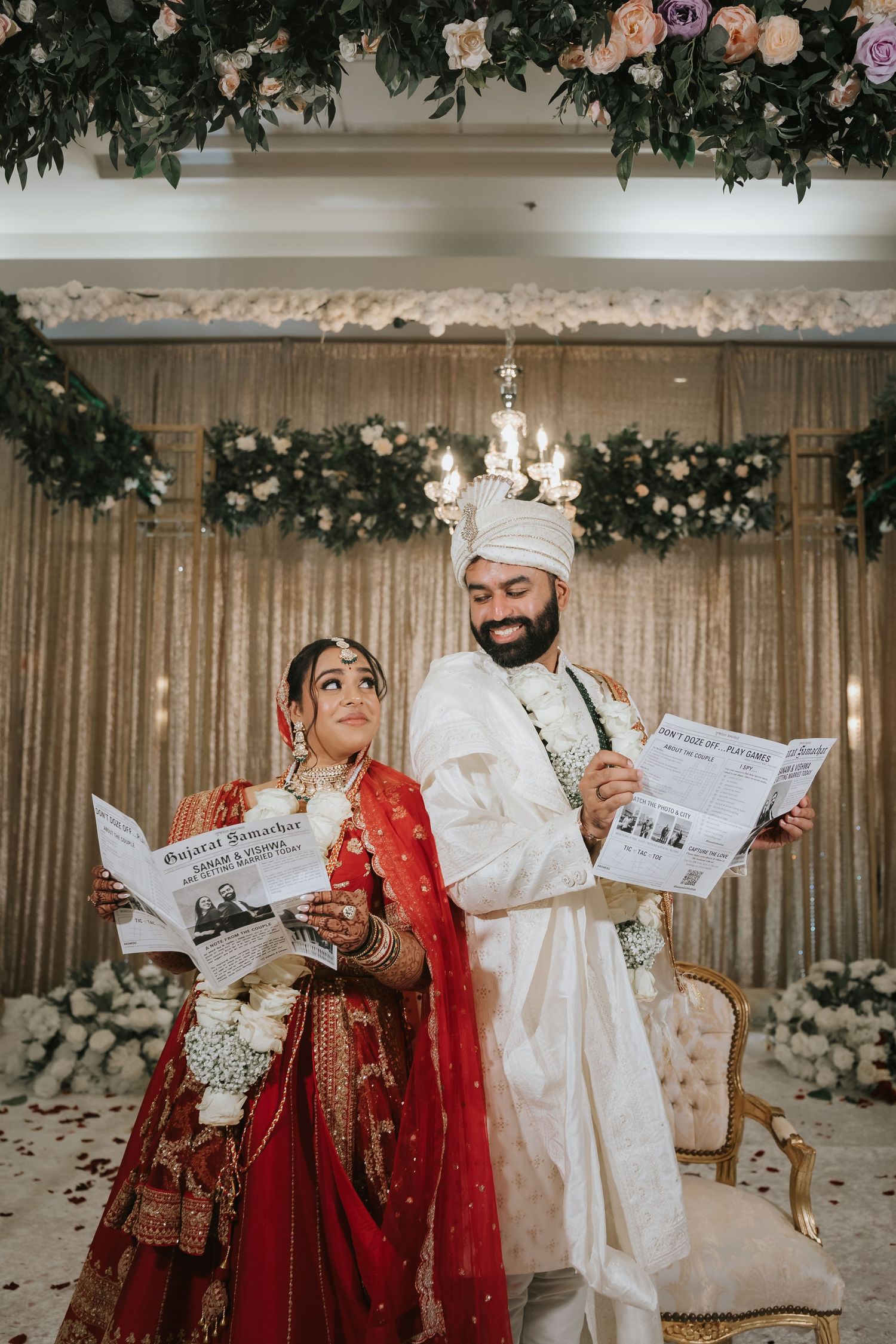 A couple in traditional Indian wedding attire reading newspapers together under floral decor at their ceremony.