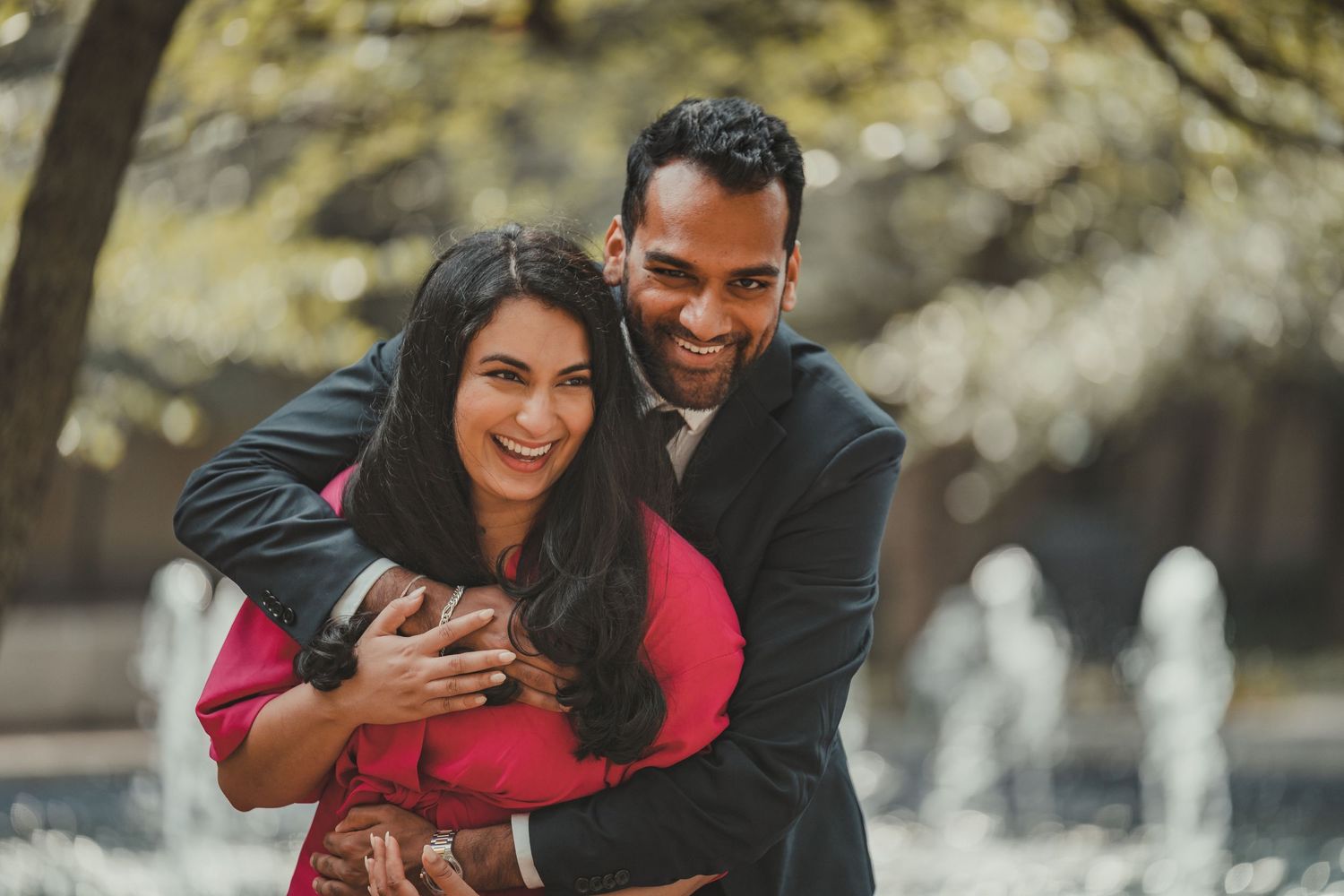 A couple shares an intimate moment in front of a fountain on a sunny day.