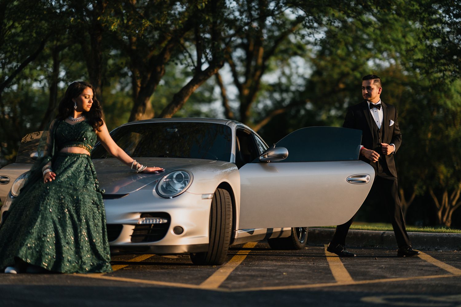 A couple poses elegantly next to a silver Porsche 911 at sunset, with trees in the background.