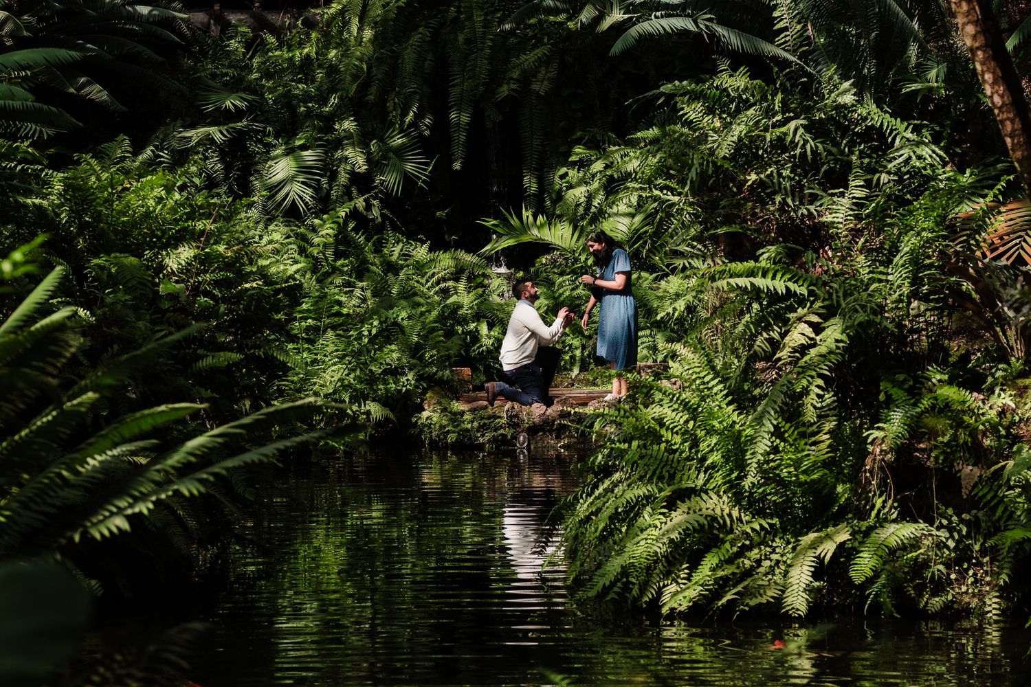 Two people sit by a serene pond surrounded by lush tropical ferns and greenery in a tranquil garden setting.