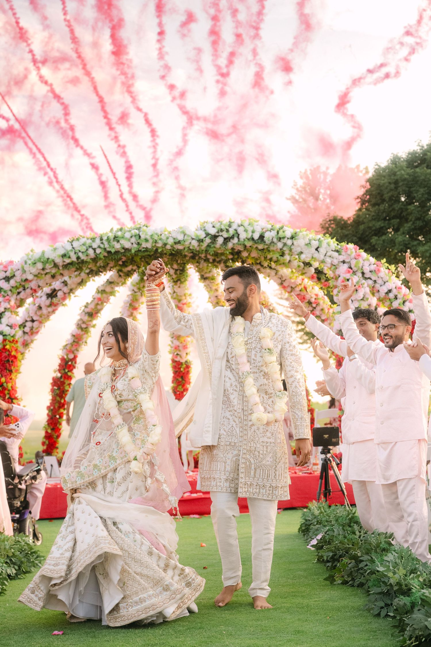Indian bride and groom celebrate wedding under floral arch with pink fireworks and guests cheering outdoors.