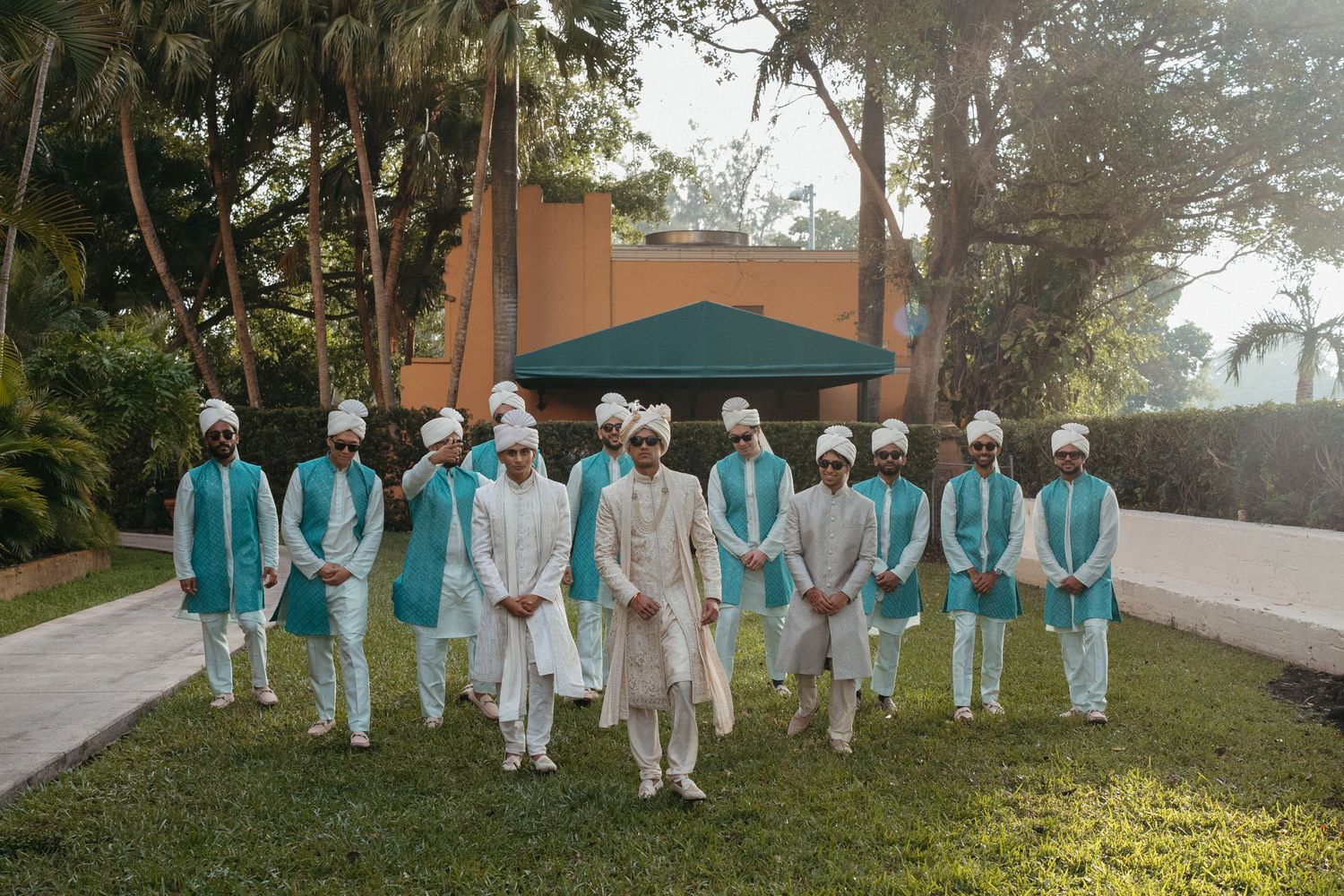 Wedding party members in coordinated turquoise and white traditional Indian attire stand together on a garden lawn.