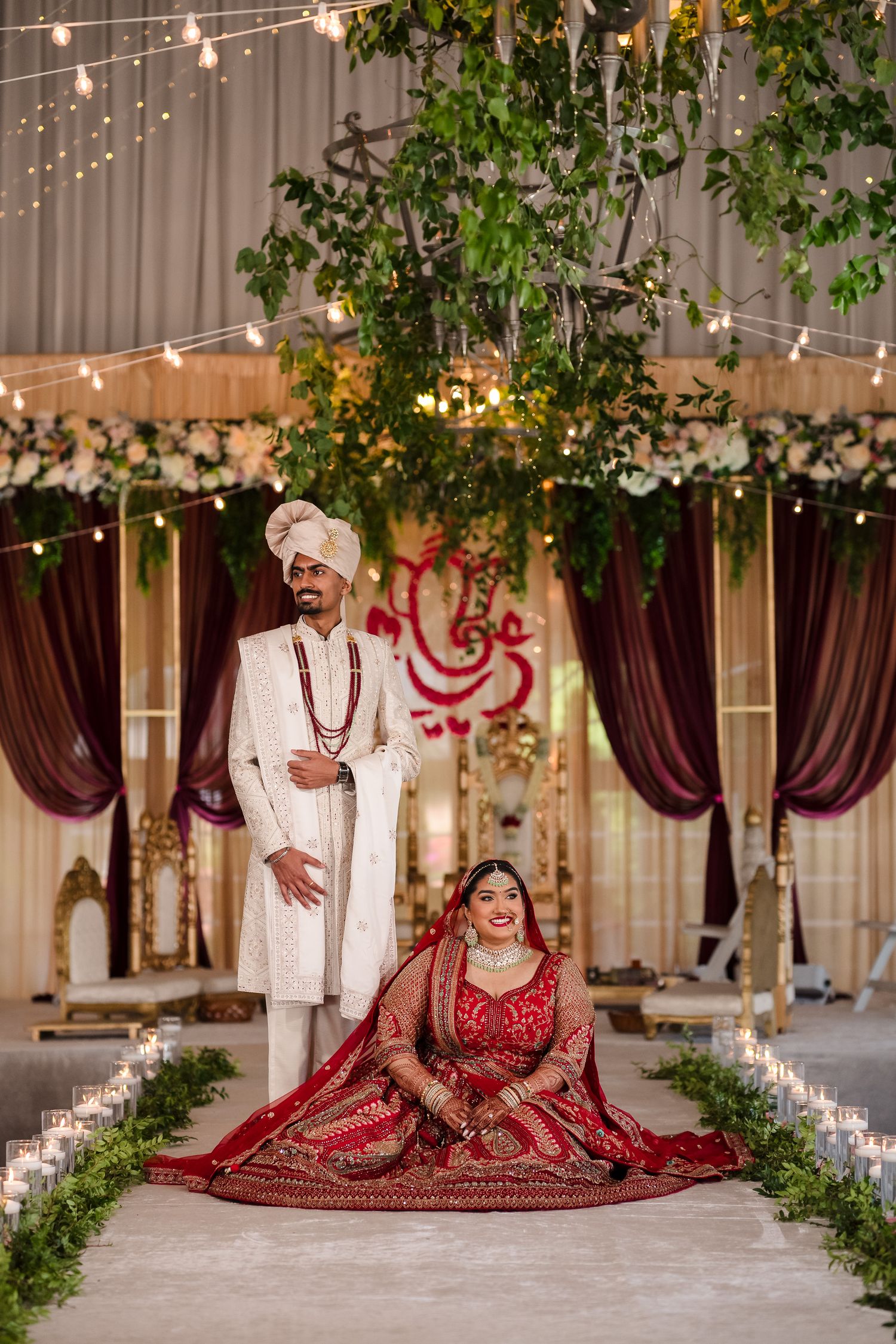 Traditional Indian wedding couple poses under decorative mandap with string lights and floral arrangements in elegant venue.