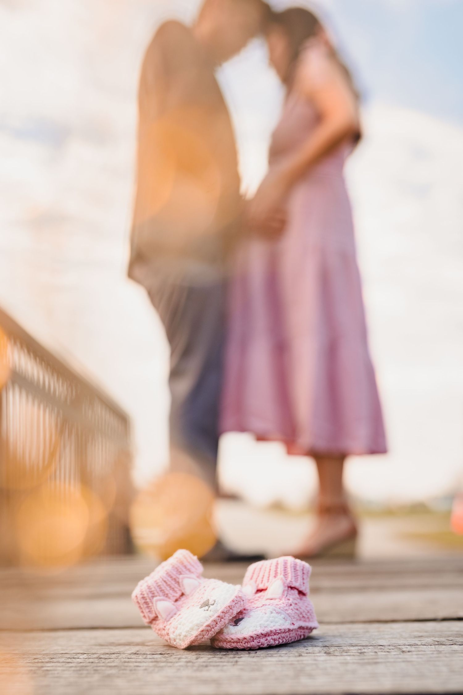 Pink knitted baby booties on wooden deck with blurred couple standing in sunlit background.