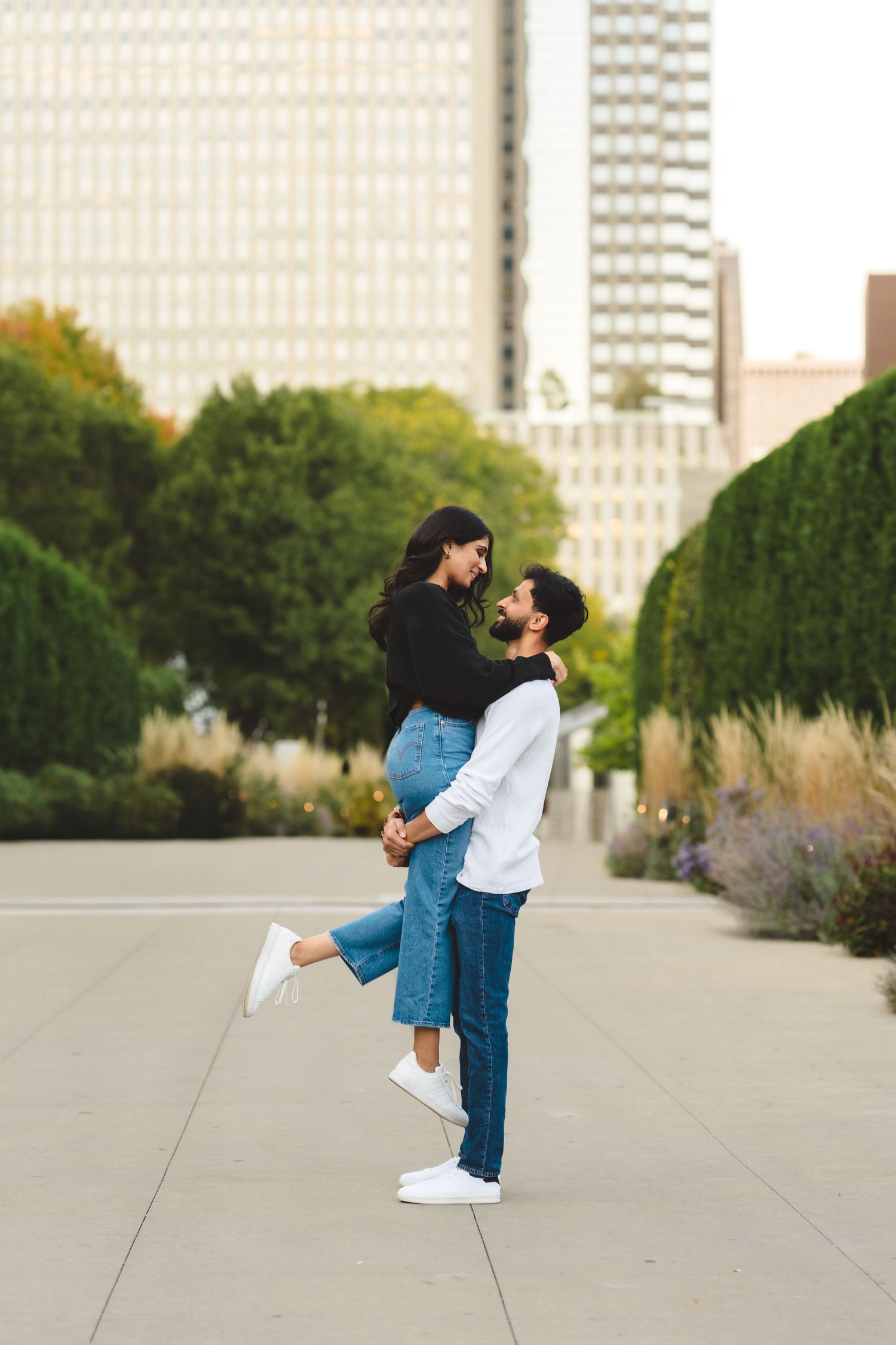 A romantic embrace between two people in an urban setting with tall buildings and landscaped gardens.