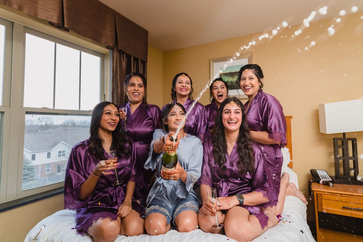 A group wearing matching purple robes celebrates with cake and champagne in a bedroom.