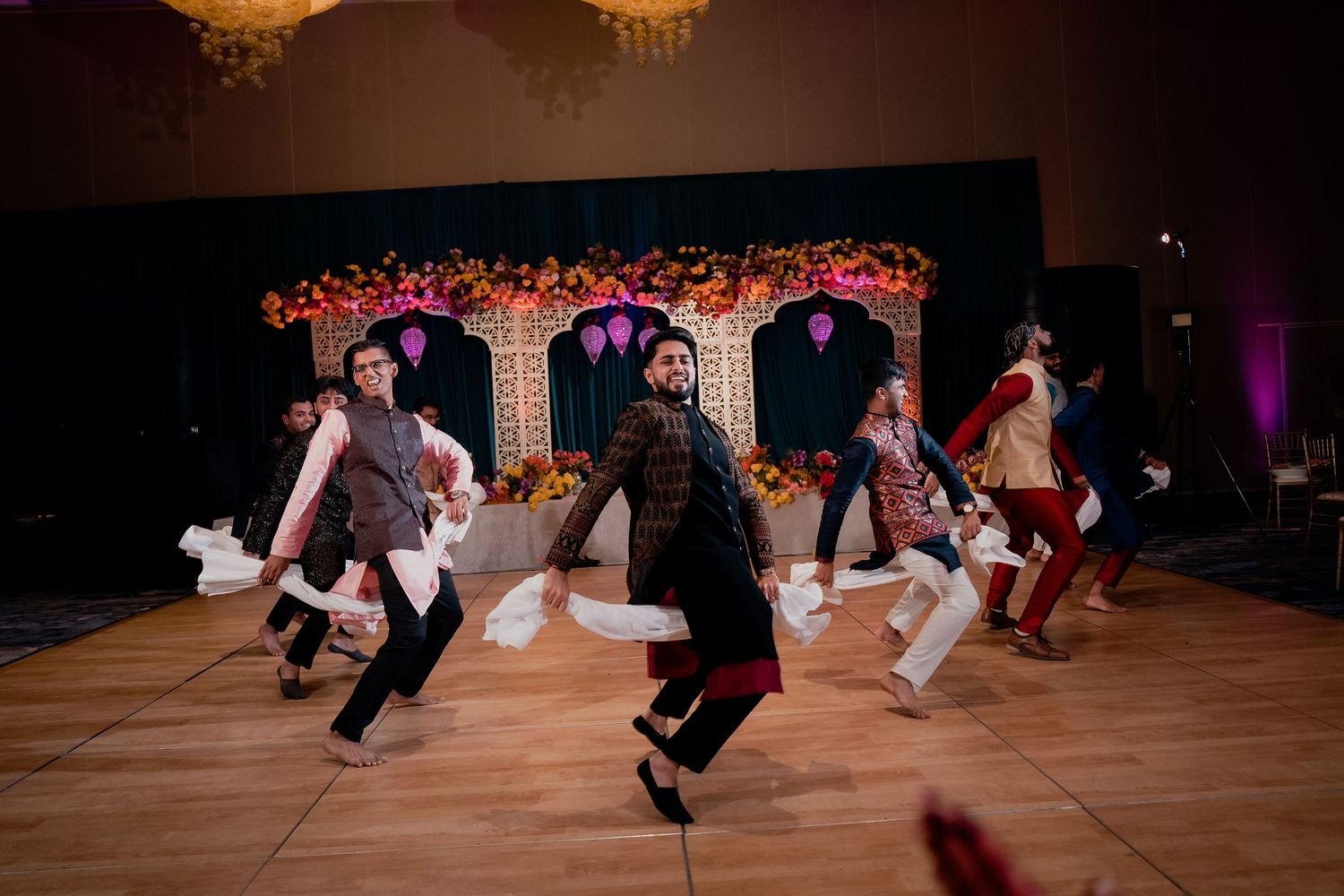 Wedding guests perform a synchronized dance routine on a ballroom floor under a floral archway at a reception.