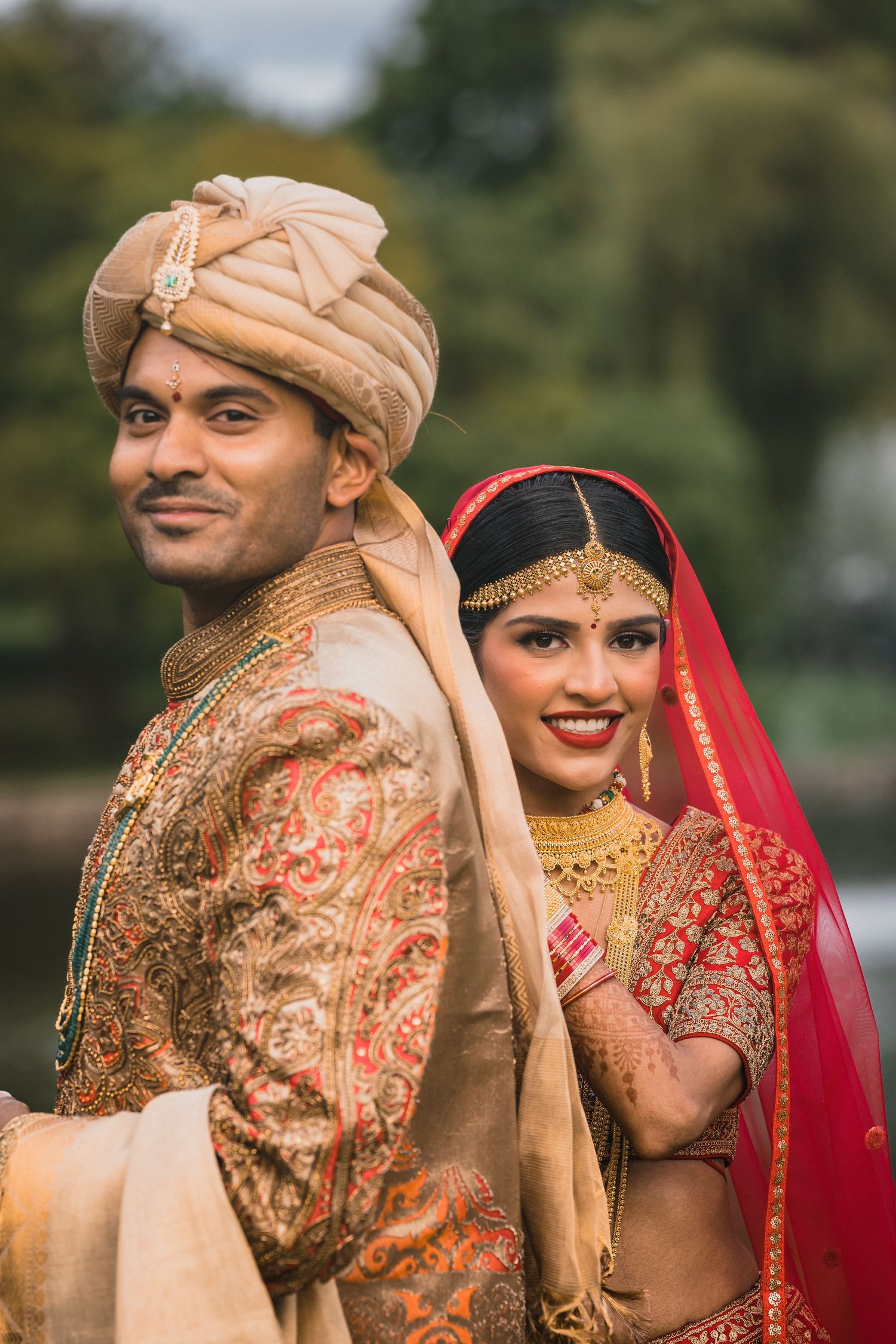 Traditional Indian wedding couple in ornate red and gold attire pose together outdoors in romantic portrait.