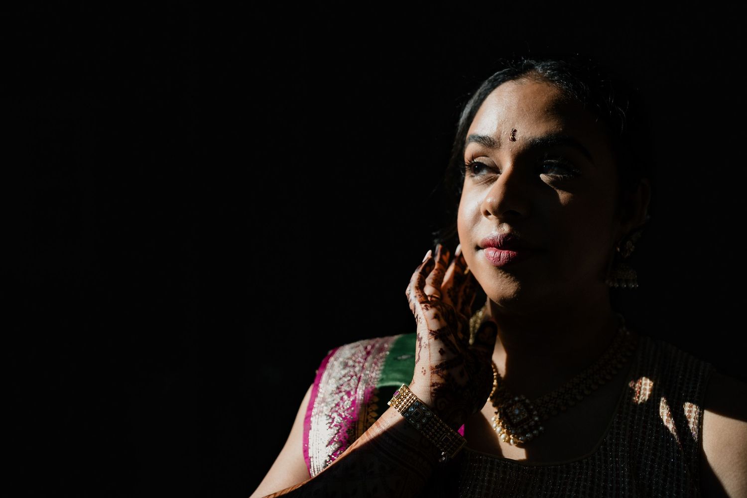 Figure in traditional South Asian attire with jewelry poses dramatically against dark background in moody lighting.