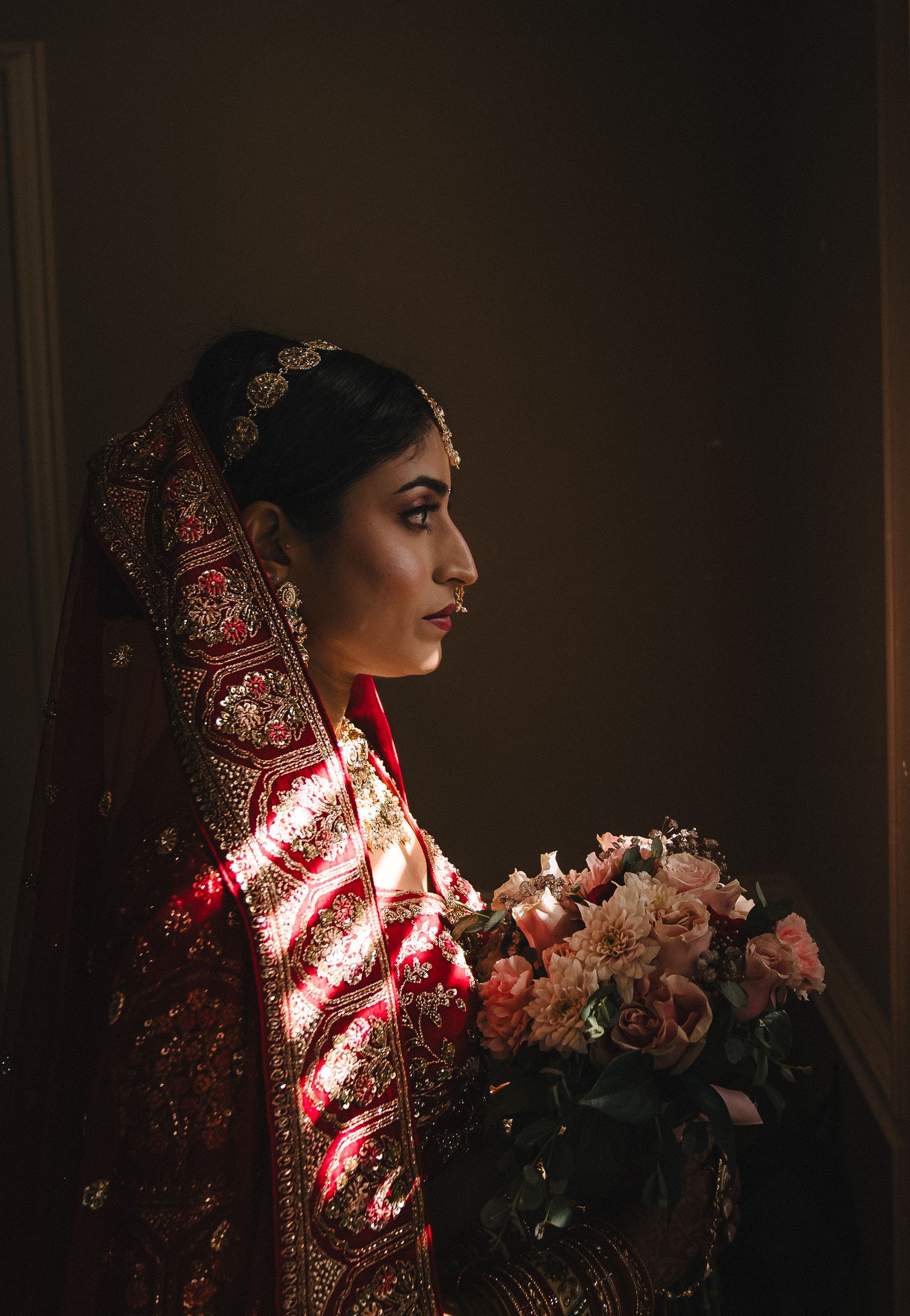 A side profile view of an Indian bride in traditional red attire holding a pink bouquet against a dark background.