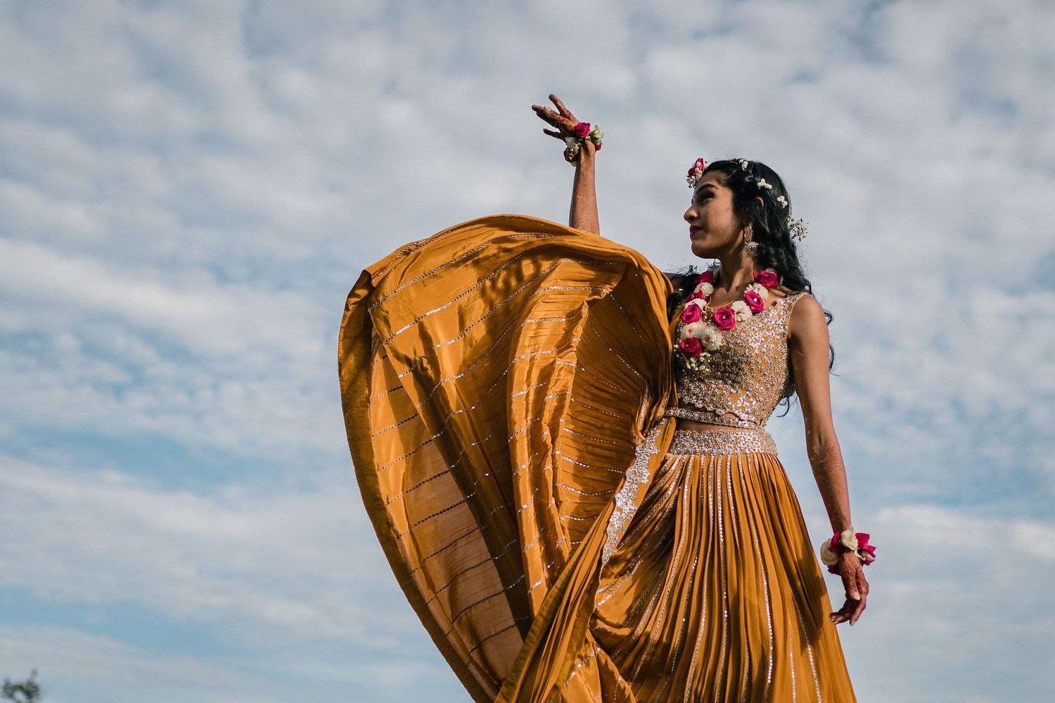 Dramatic fashion pose with flowing gold dress against cloudy sky.