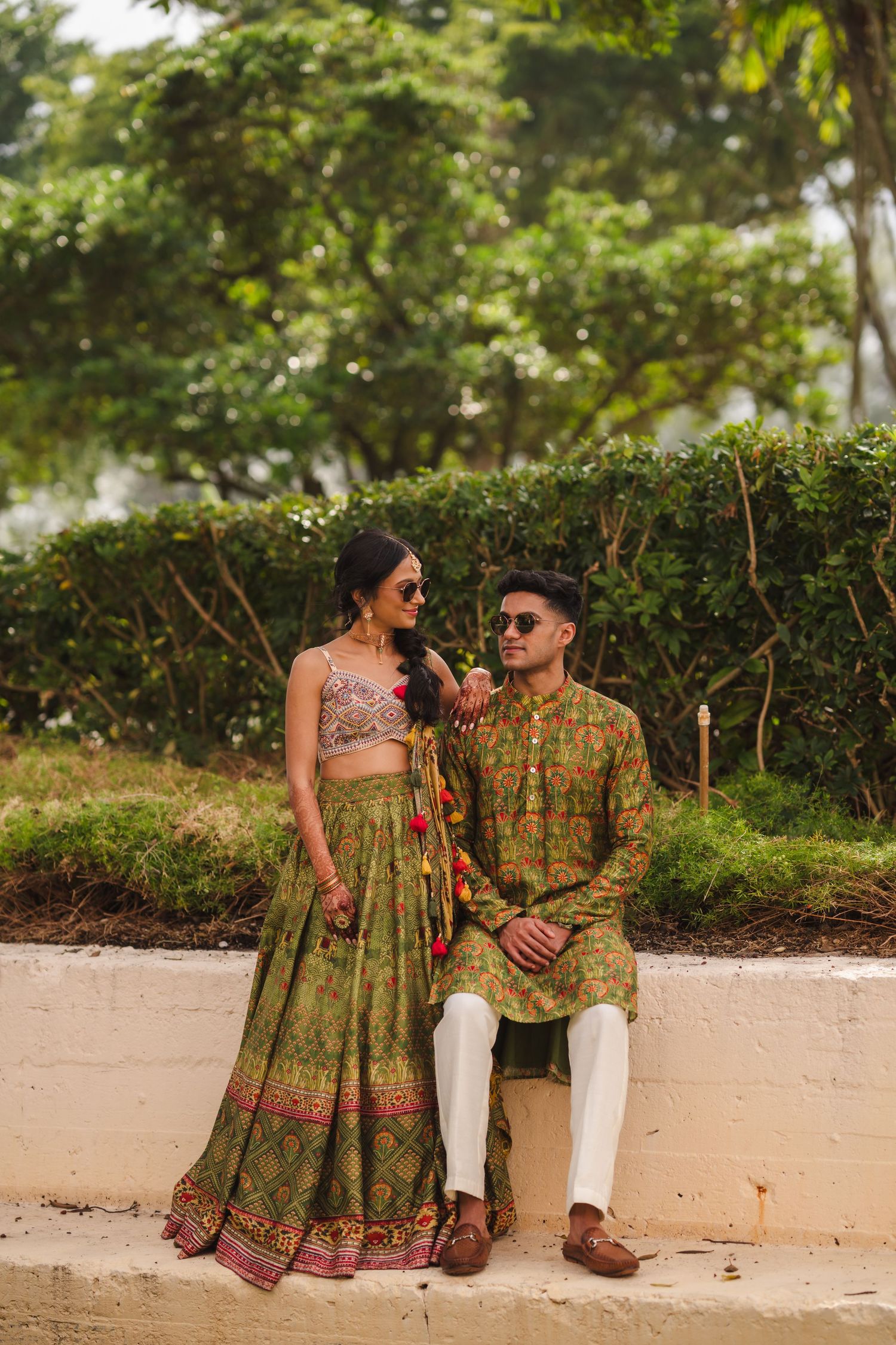 Traditional Indian couple wearing elegant ethnic outfits pose together against lush garden background.