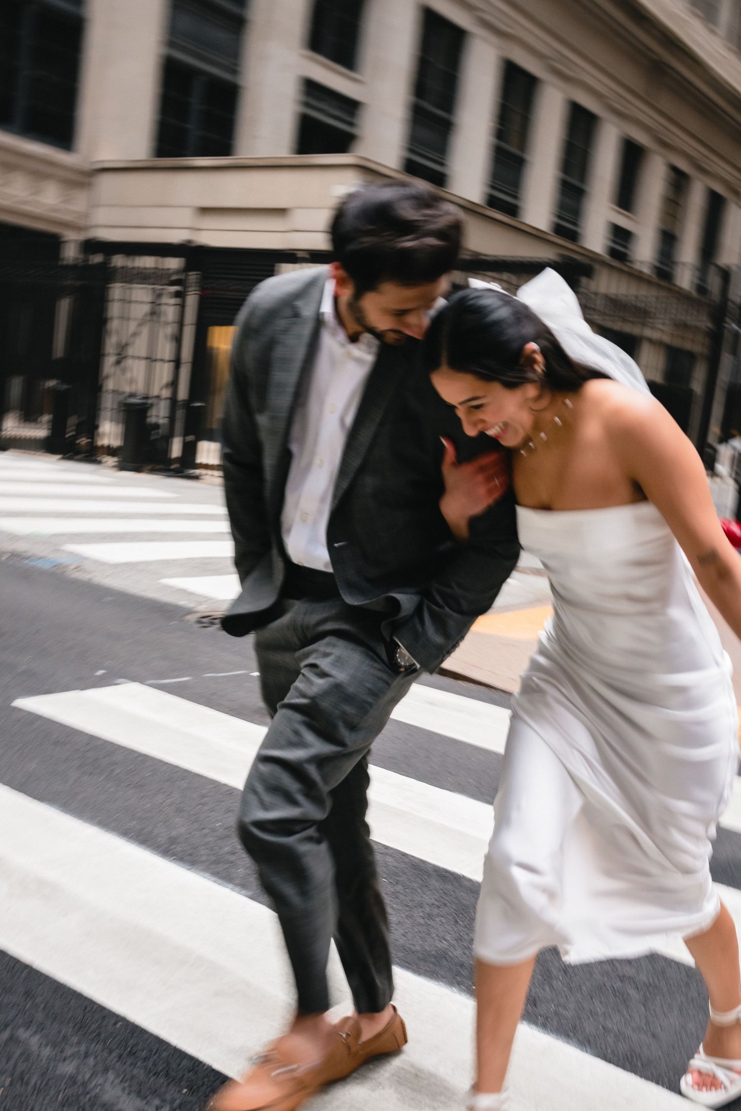 A couple in wedding attire playfully crosses an urban crosswalk together on their special day.