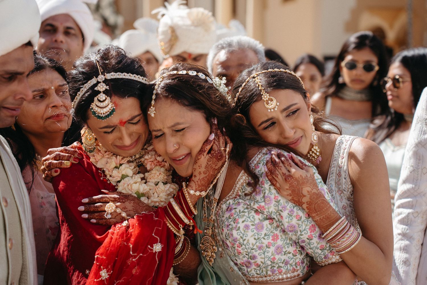 Three people embrace emotionally during a traditional South Asian wedding ceremony with guests looking on in the background.