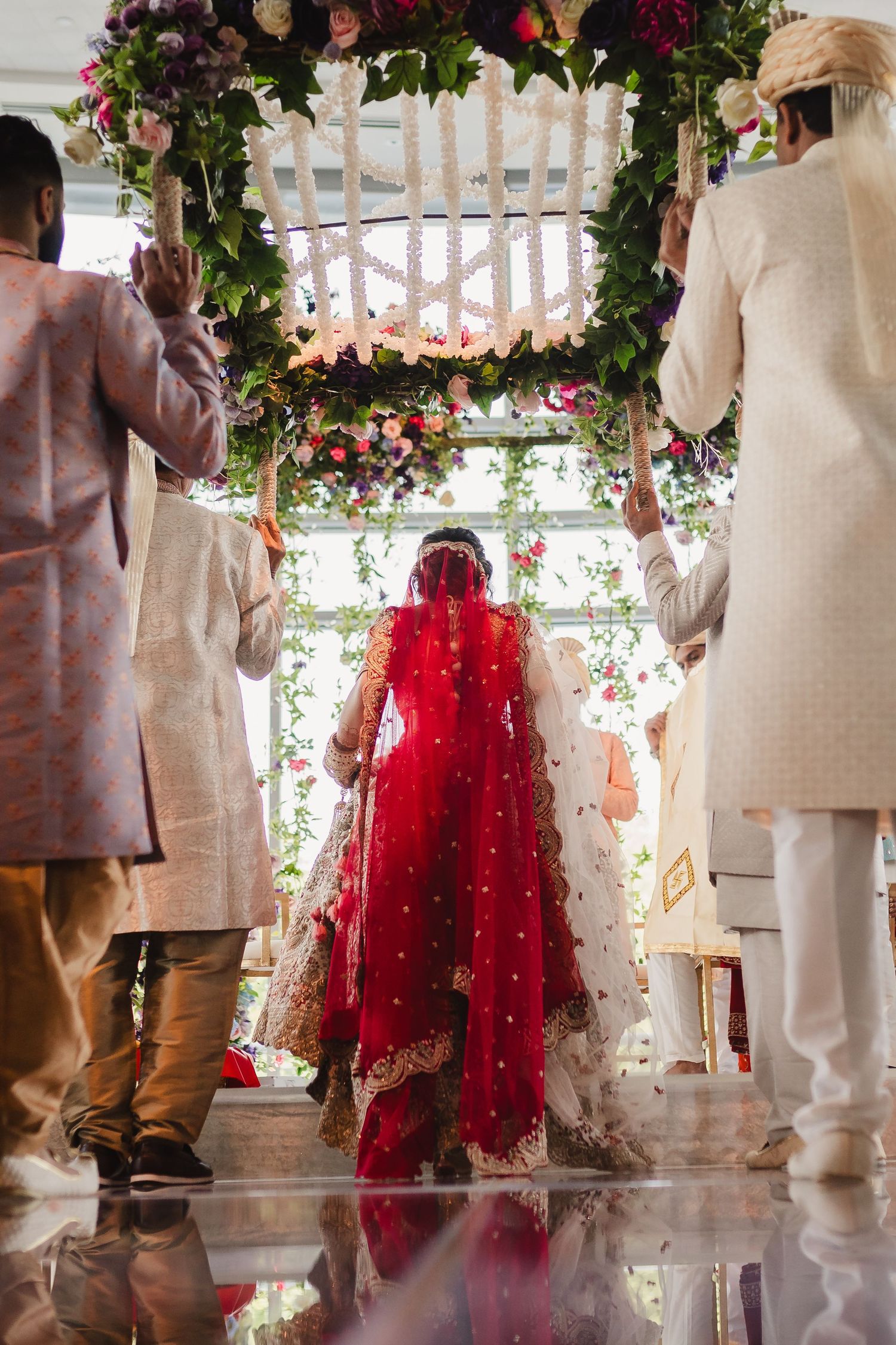 Traditional wedding ceremony under a floral archway decorated with roses and greenery, reflected in polished marble floor.