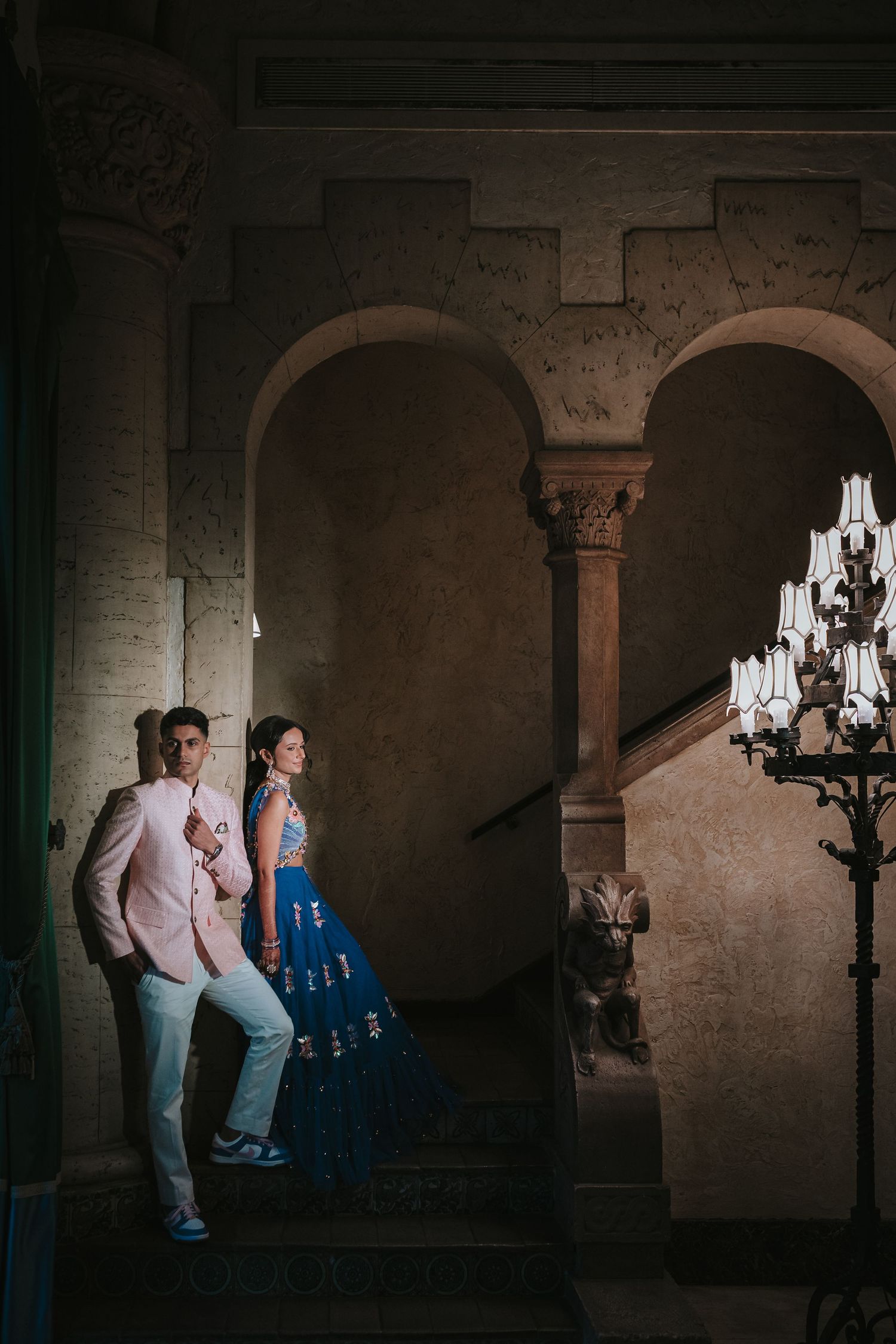 A couple poses on stone stairs near an illuminated chandelier in an elegant architectural setting.