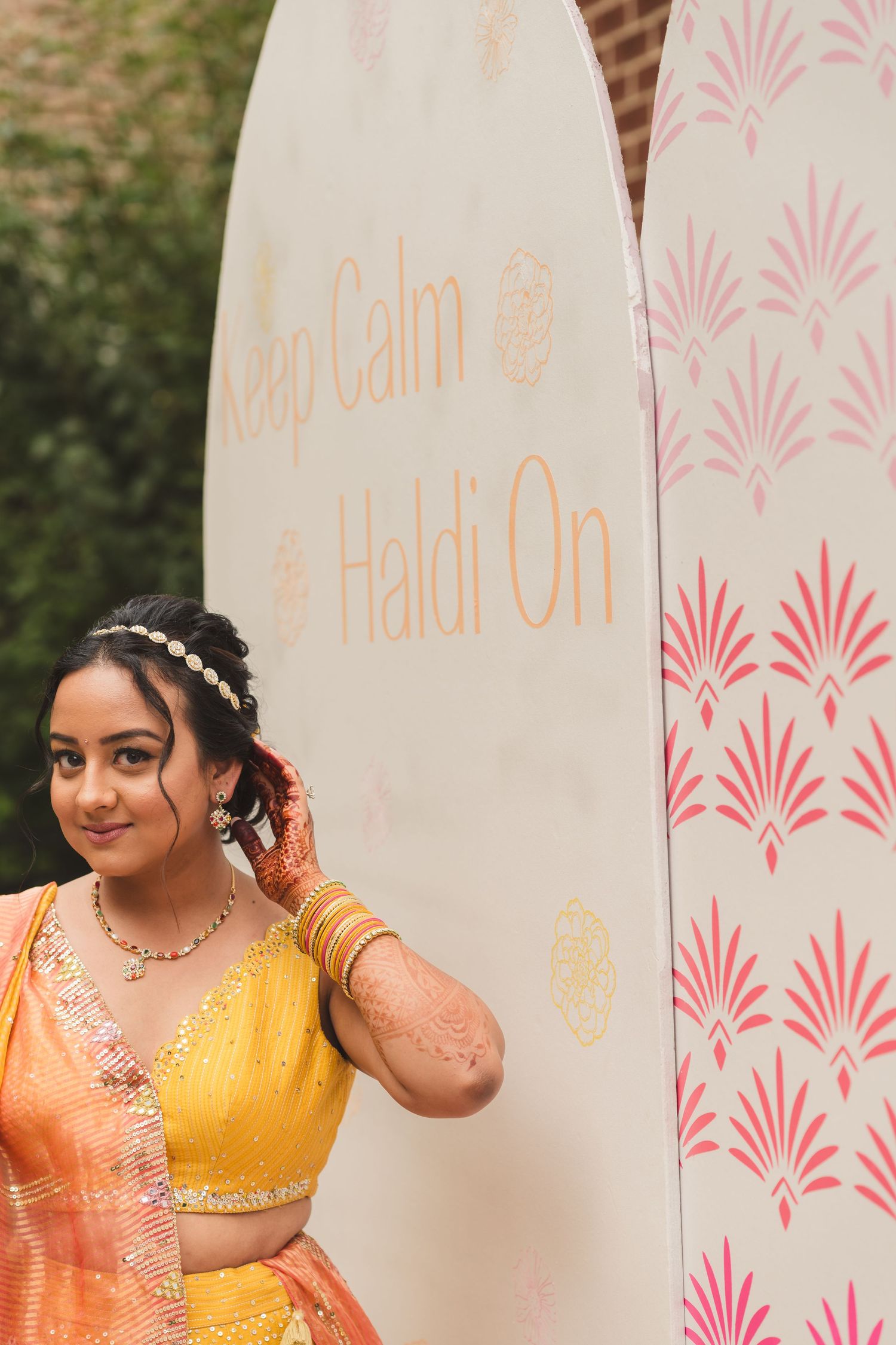 Bride in yellow lehenga poses next to decorative wall with text.