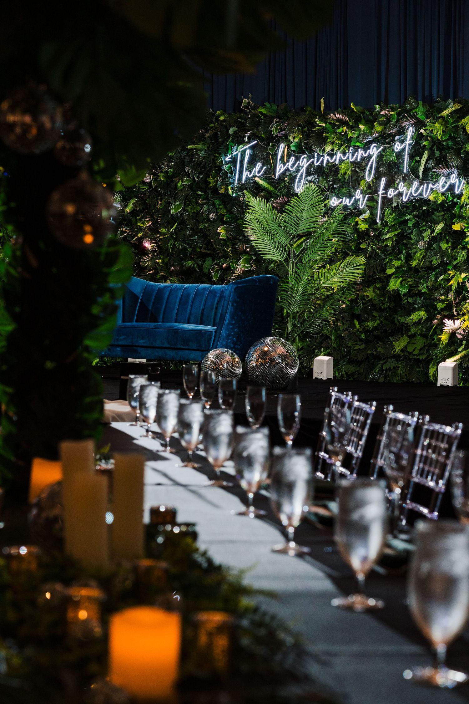 Elegant wedding reception setup with blue velvet seating and lush greenery wall backdrop.