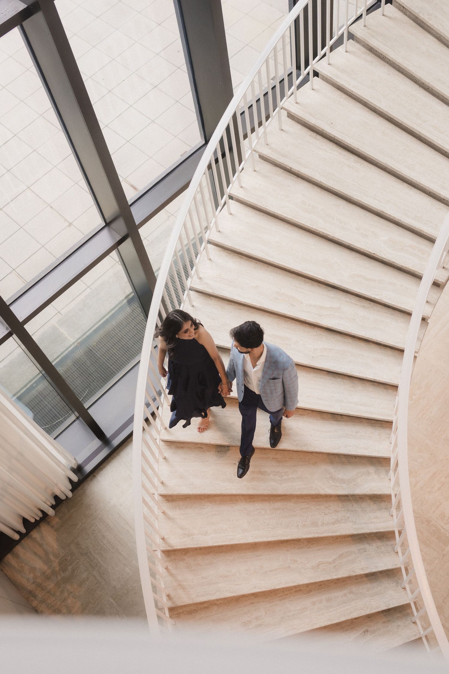 Two people stand together on a modern white spiral staircase near glass windows in a minimalist interior space.