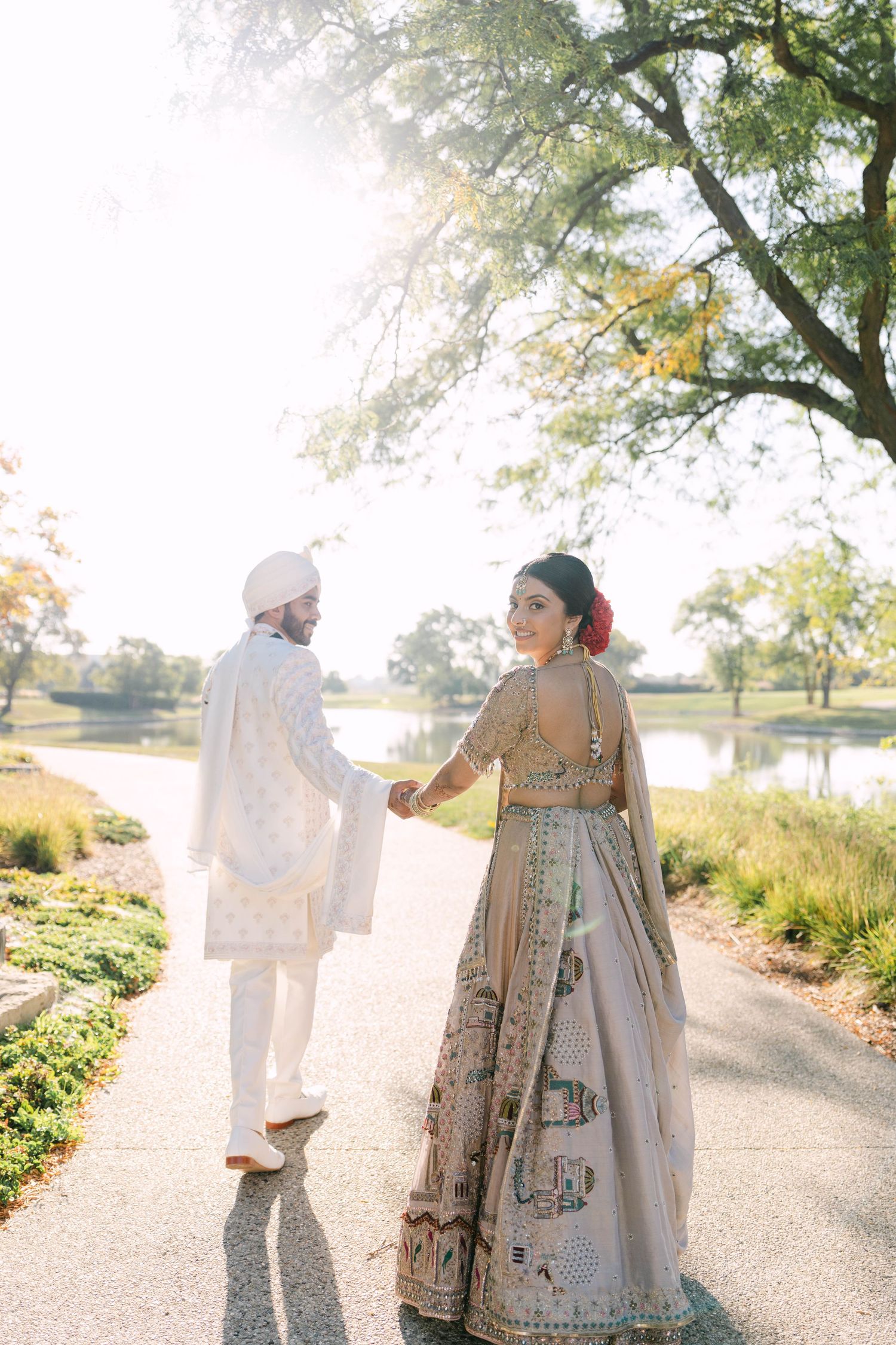 A couple in traditional Indian wedding attire walking together on a sunlit path surrounded by greenery.