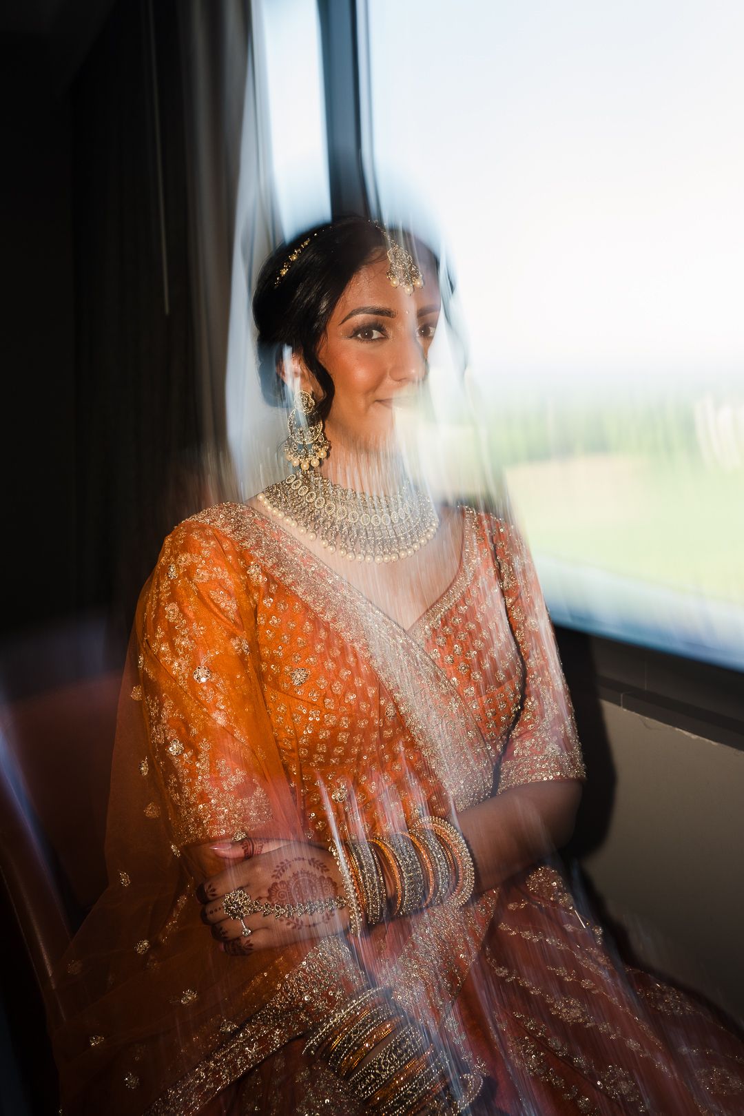 A bride in a glittering peach lehenga and pearl jewelry sits by a window in soft natural light.