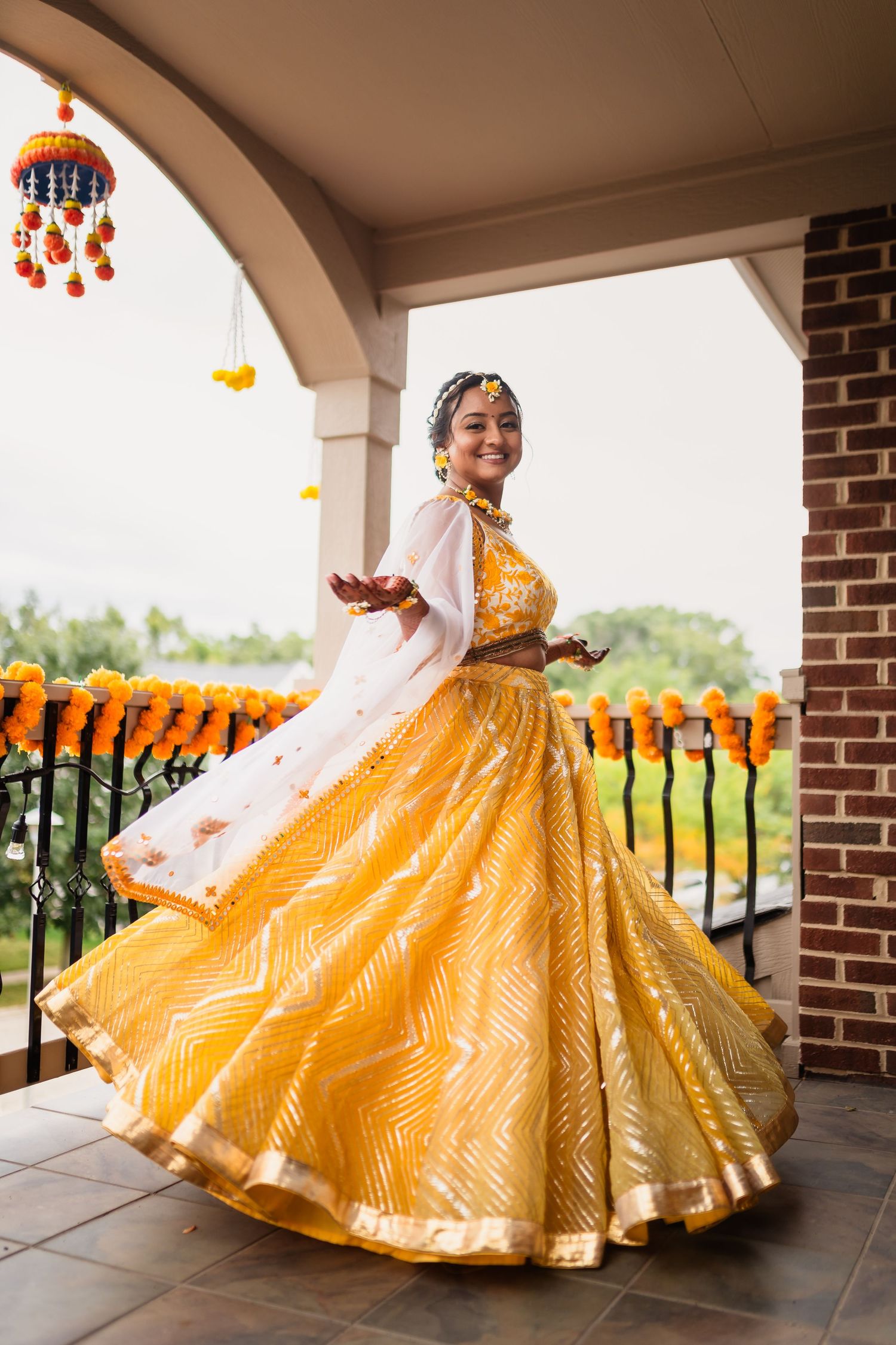 A yellow lehenga twirls gracefully on a porch decorated with marigold garlands and hanging lanterns.