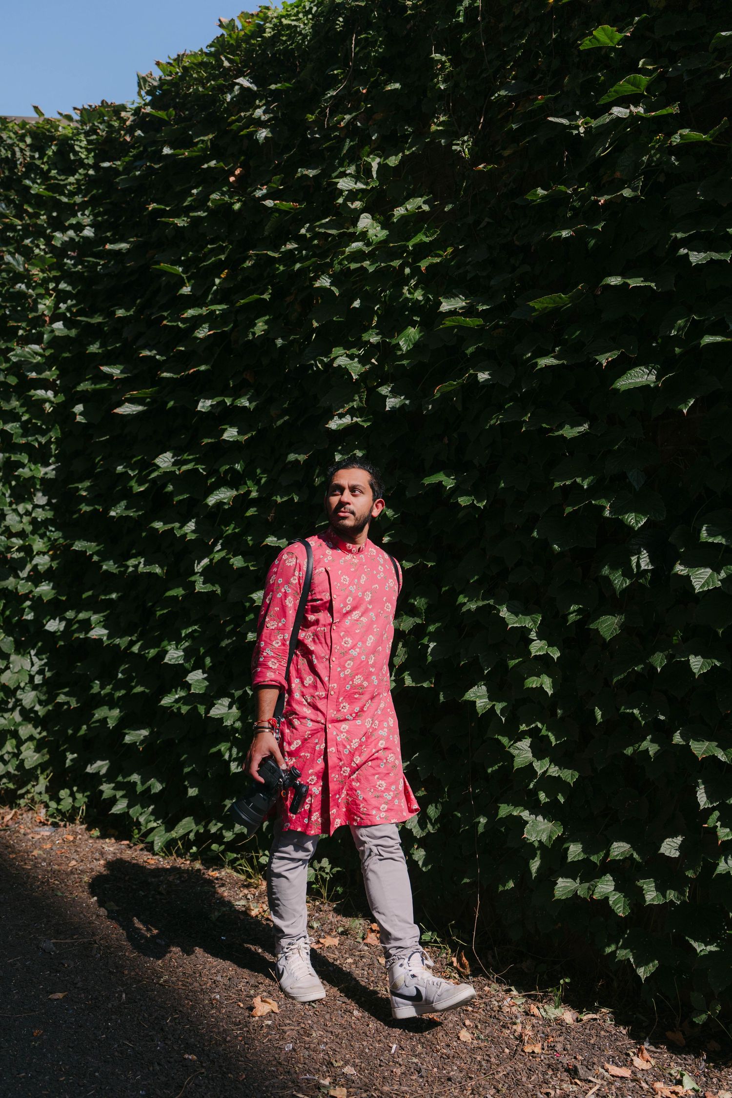 A person in a pink polka dot shirt and gray pants stands against a backdrop of lush green foliage.