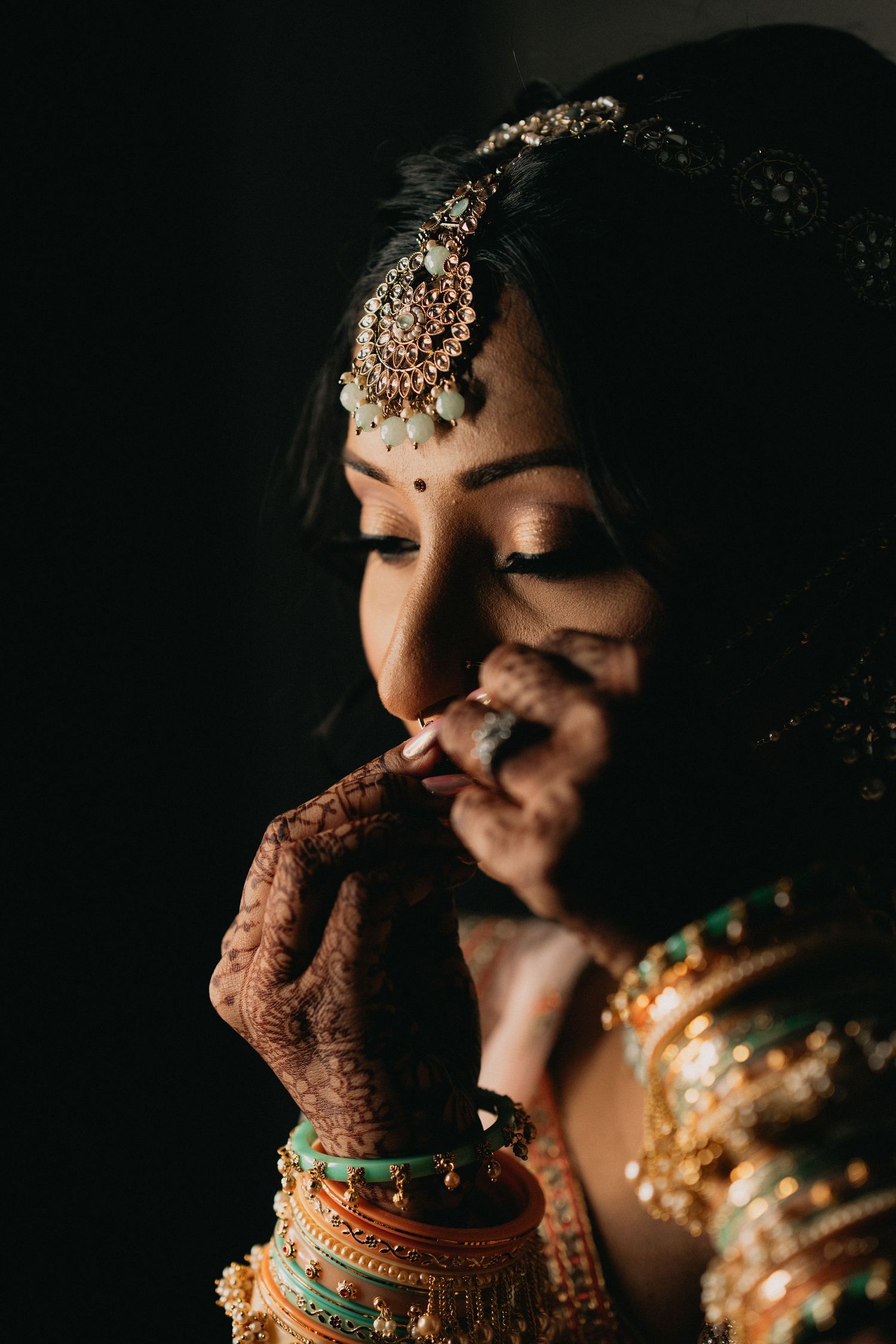 Traditional Indian bride wearing ornate jewelry and henna in dramatic lighting with dark background.