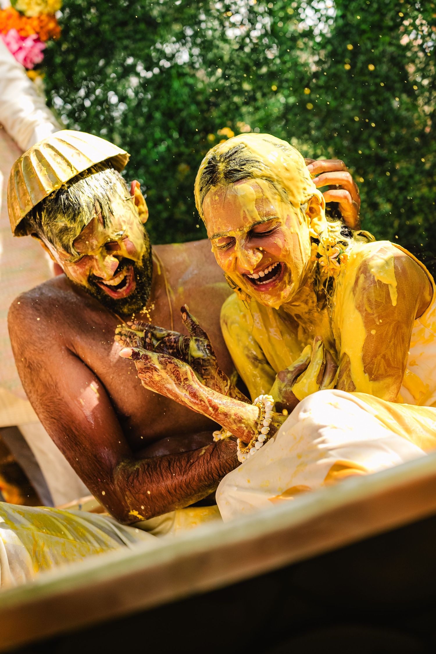 Two people covered in yellow powder laugh joyfully during what appears to be a Holi festival celebration outdoors.