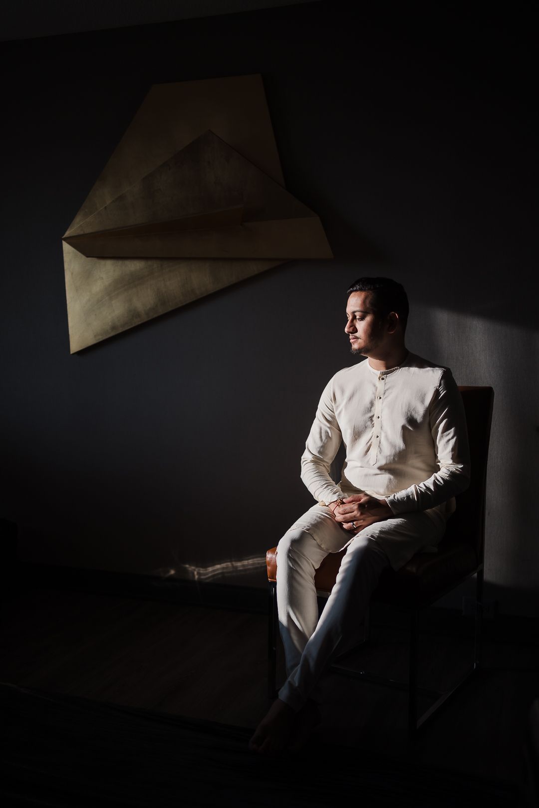Moody studio portrait of figure in white sweater seated against dark wall with geometric paper art installation.