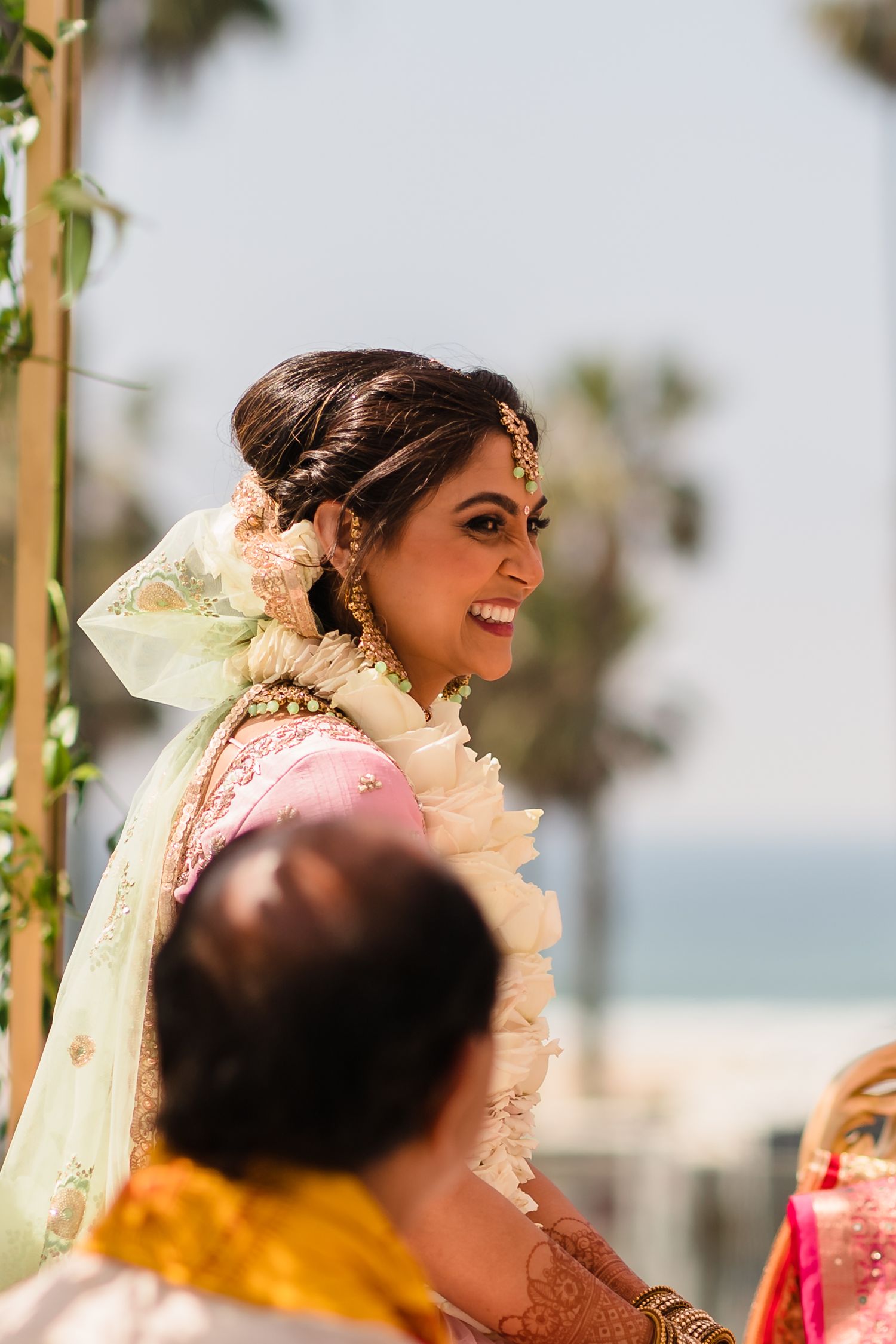 A traditional Indian bride in pink and cream attire smiling during a beachside wedding ceremony.