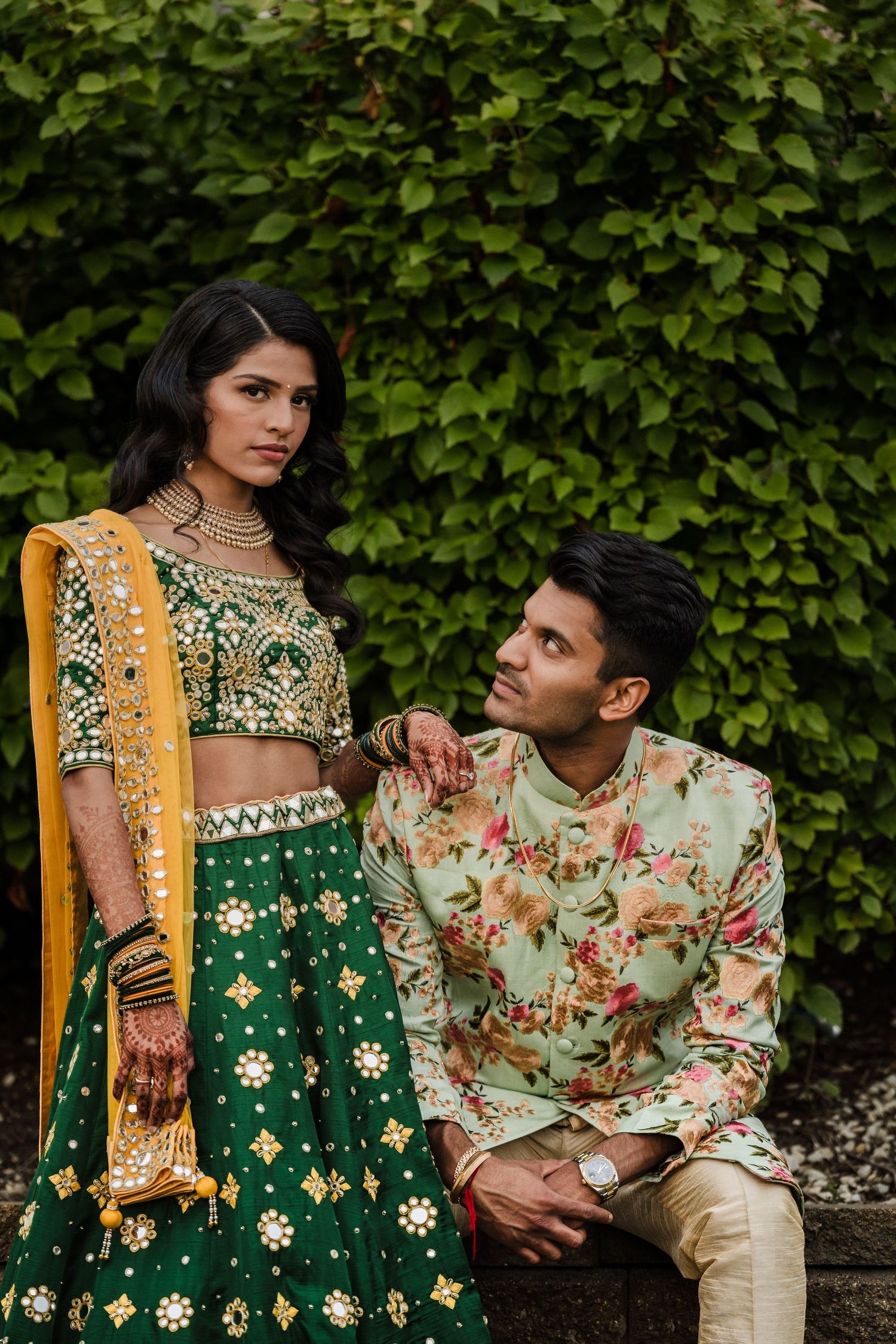 Couple in traditional Indian wedding attire pose against lush green garden backdrop.