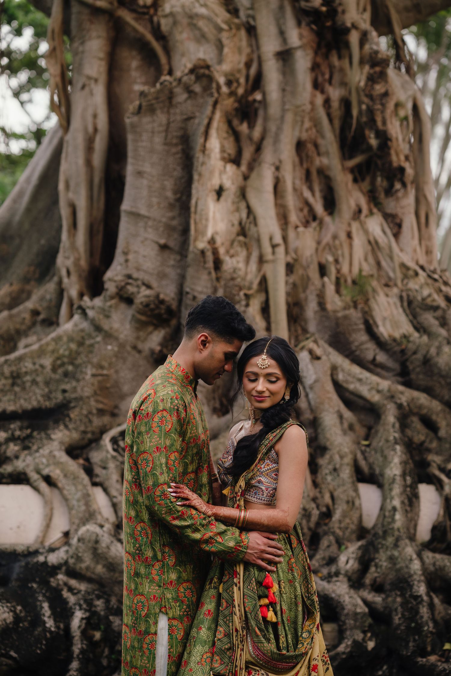 A couple in green traditional Indian outfits embrace near a large banyan tree with exposed roots.