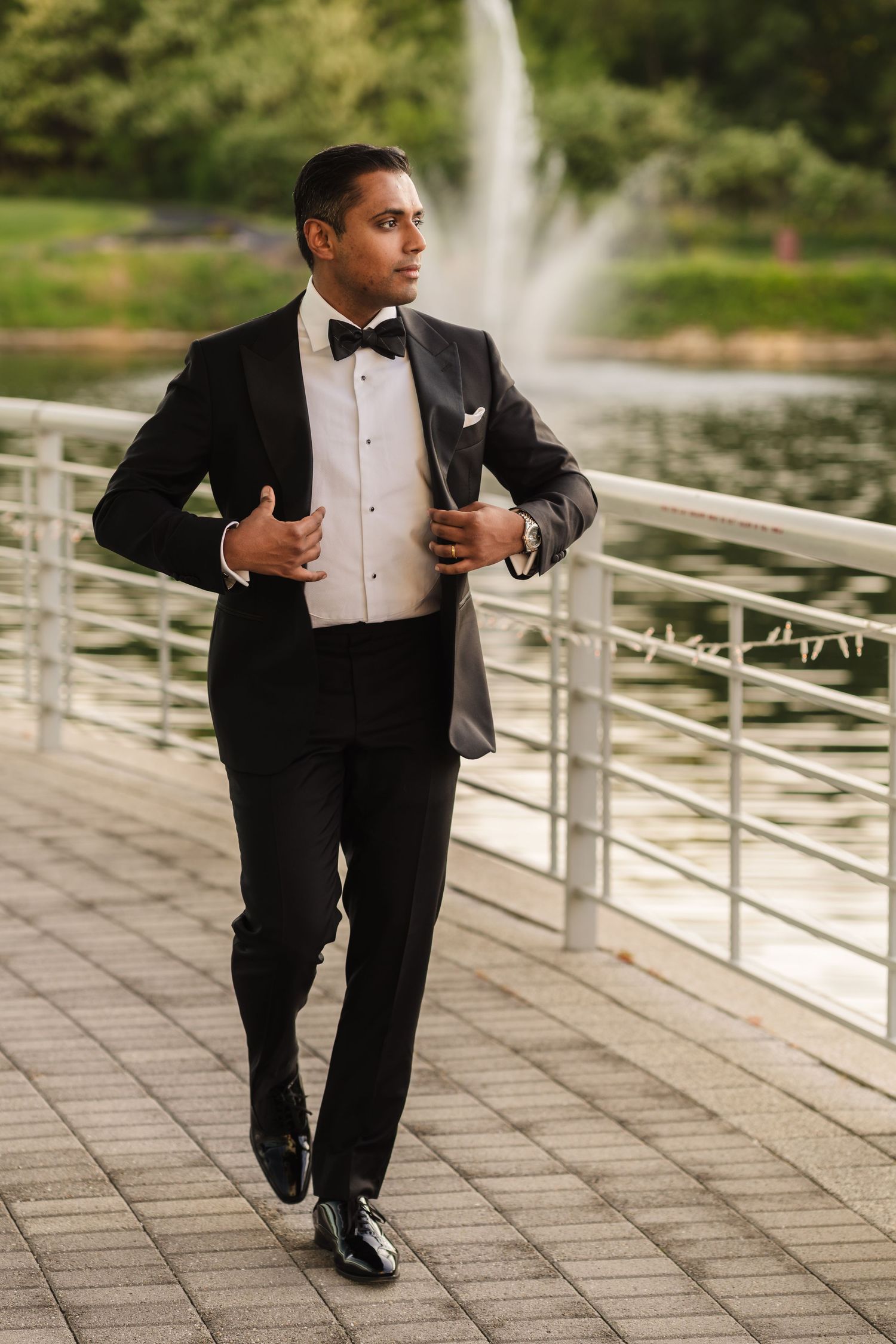 Groom in classic black tuxedo poses on waterfront deck with fountain in background.