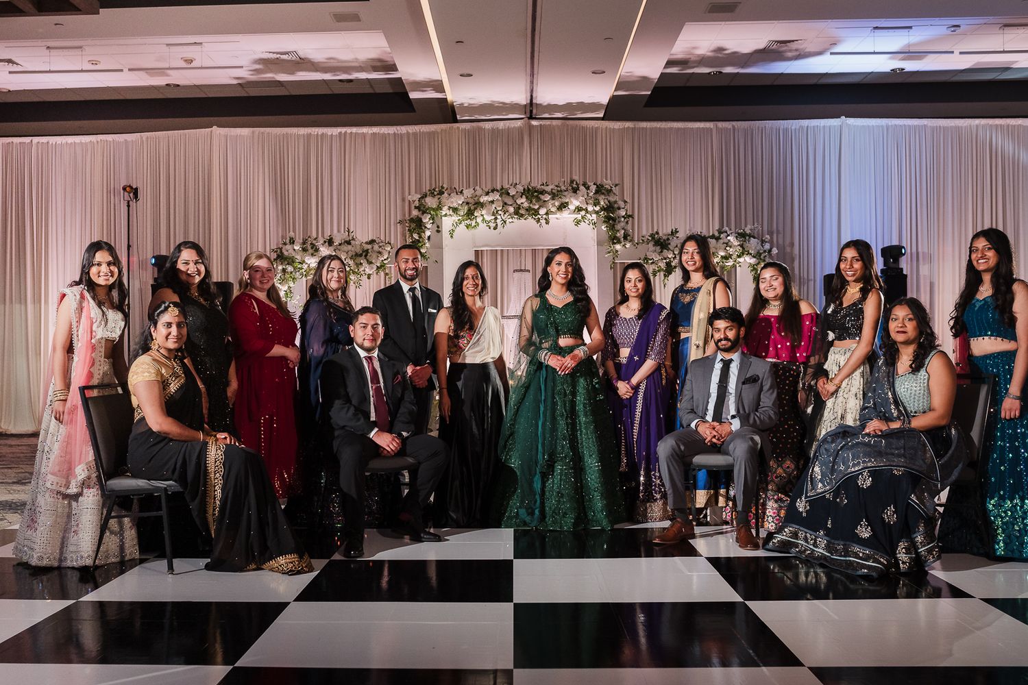 A group photo at a formal event with guests in elegant attire gathered in front of a floral arch on a black and white checkered floor.
