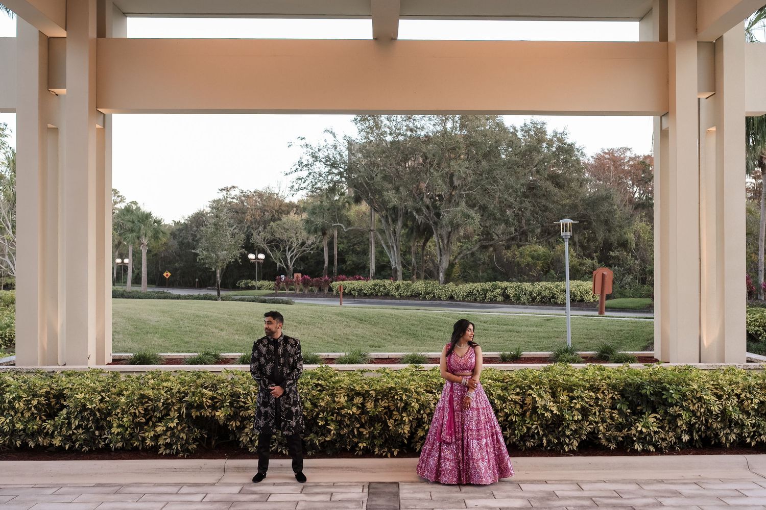 Two children in formal attire pose apart under a covered walkway with landscaped grounds in the background.