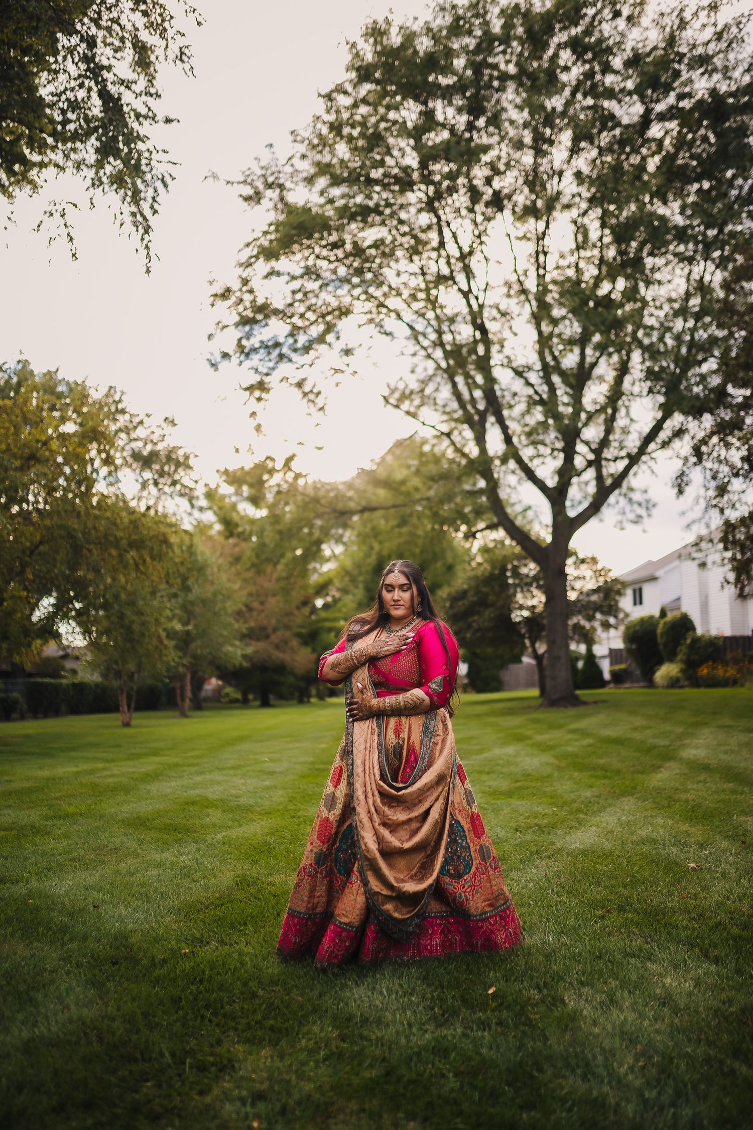 A person wearing an elegant pink and gold traditional lehenga stands on a manicured lawn surrounded by trees at dusk.